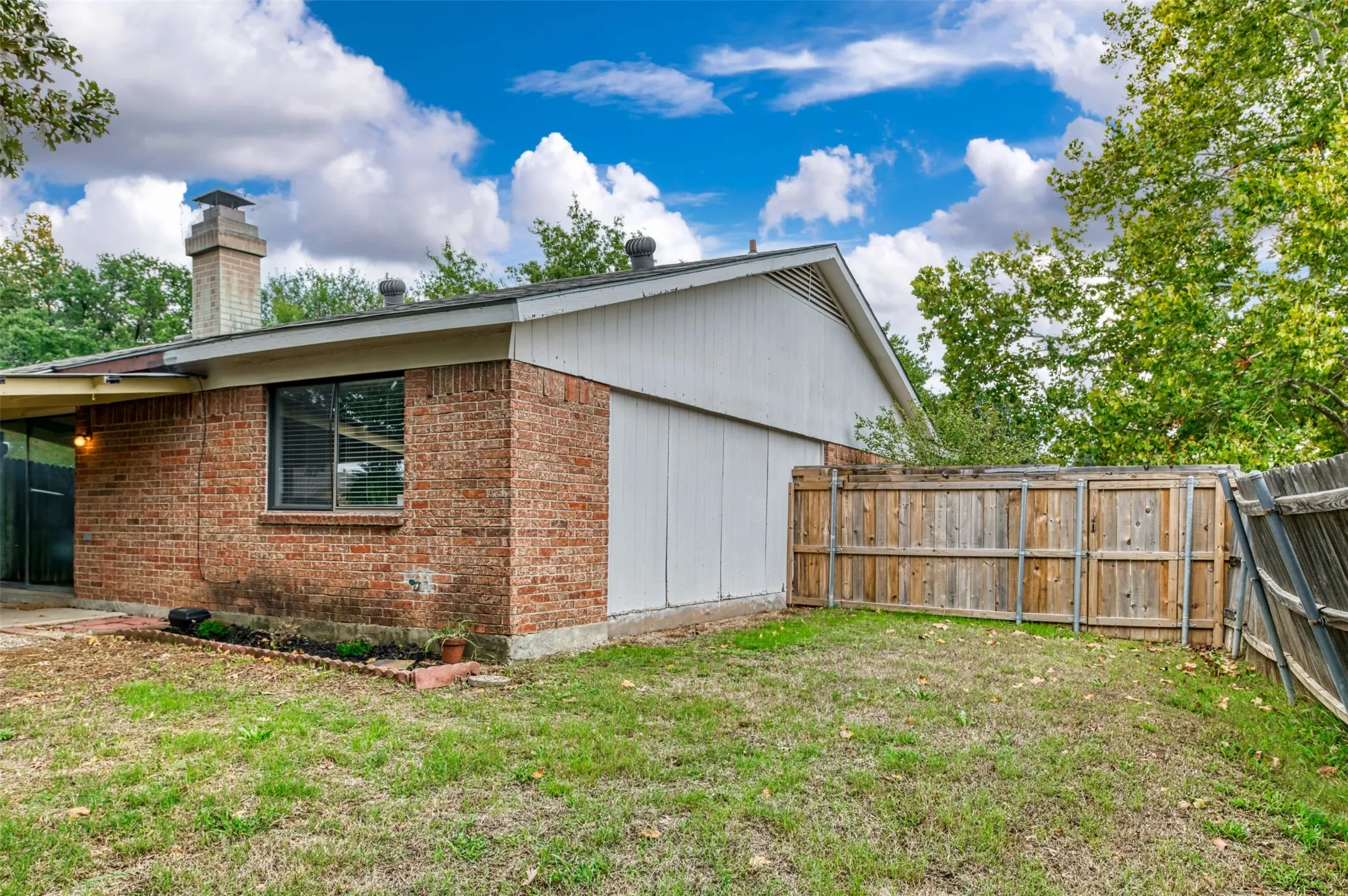Brick exterior with a prominent chimney, a window with blinds, and a wooden privacy fence enclosing a grassy area
