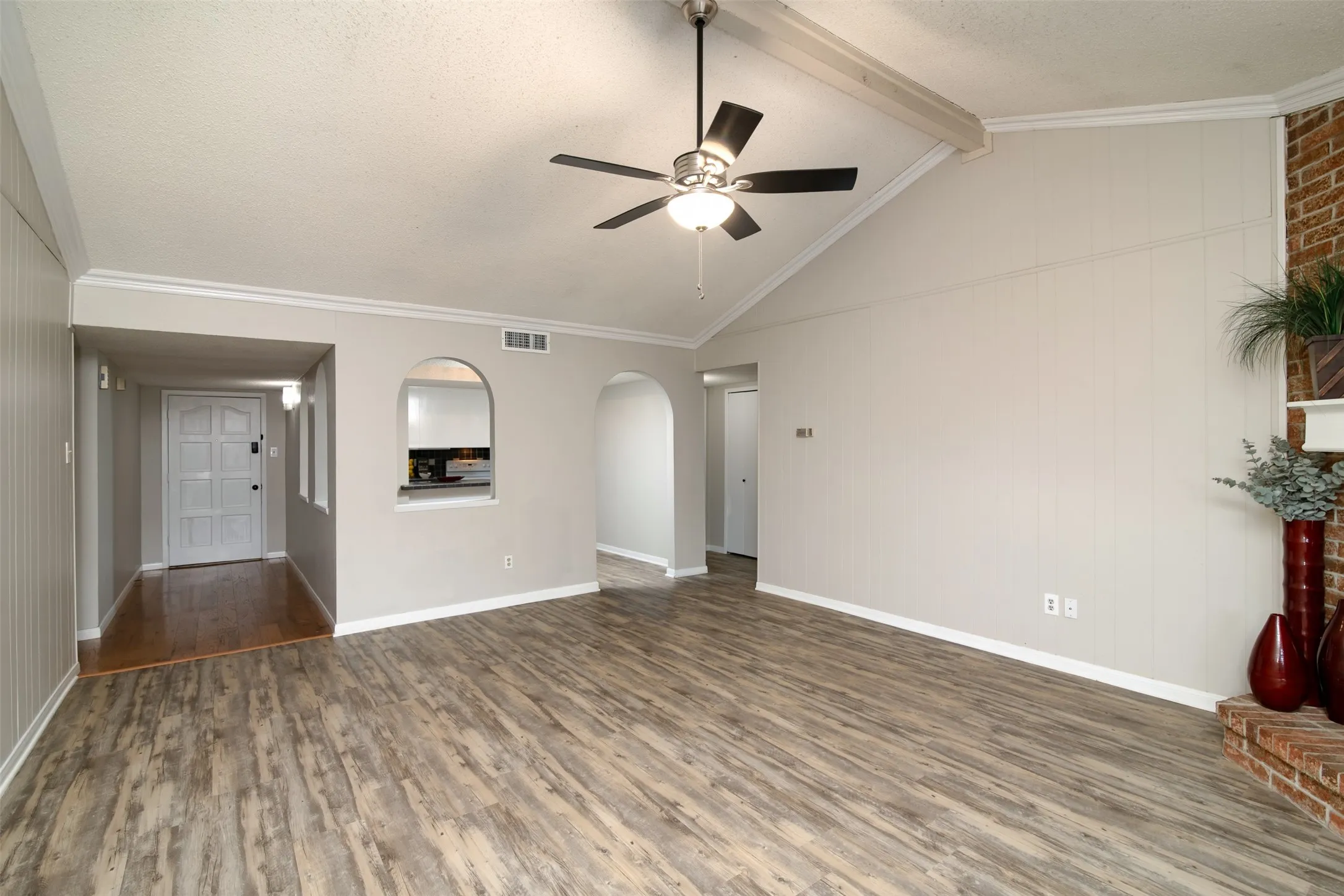 Vaulted ceiling with exposed beam, ceiling fan, brick-faced fireplace, wood-finish flooring, and arched architectural openings