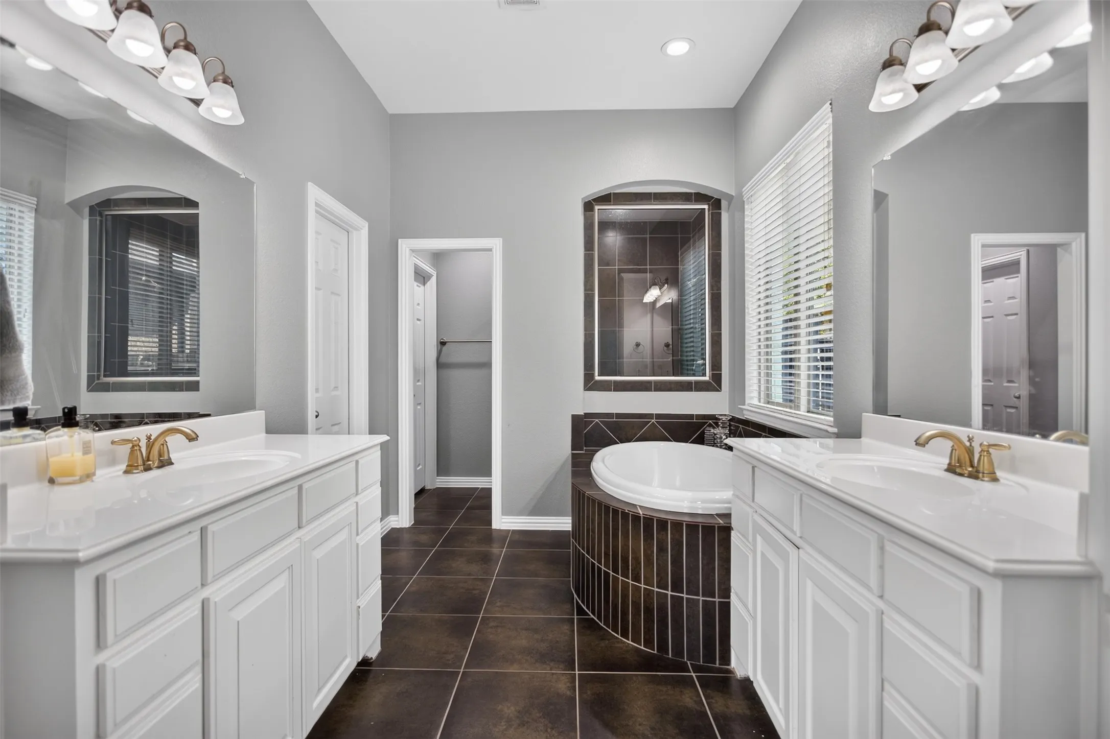 Dual vanity with white cabinetry and integrated sinks, large soaking tub, arched wall mirror, tile flooring, and a window with blinds