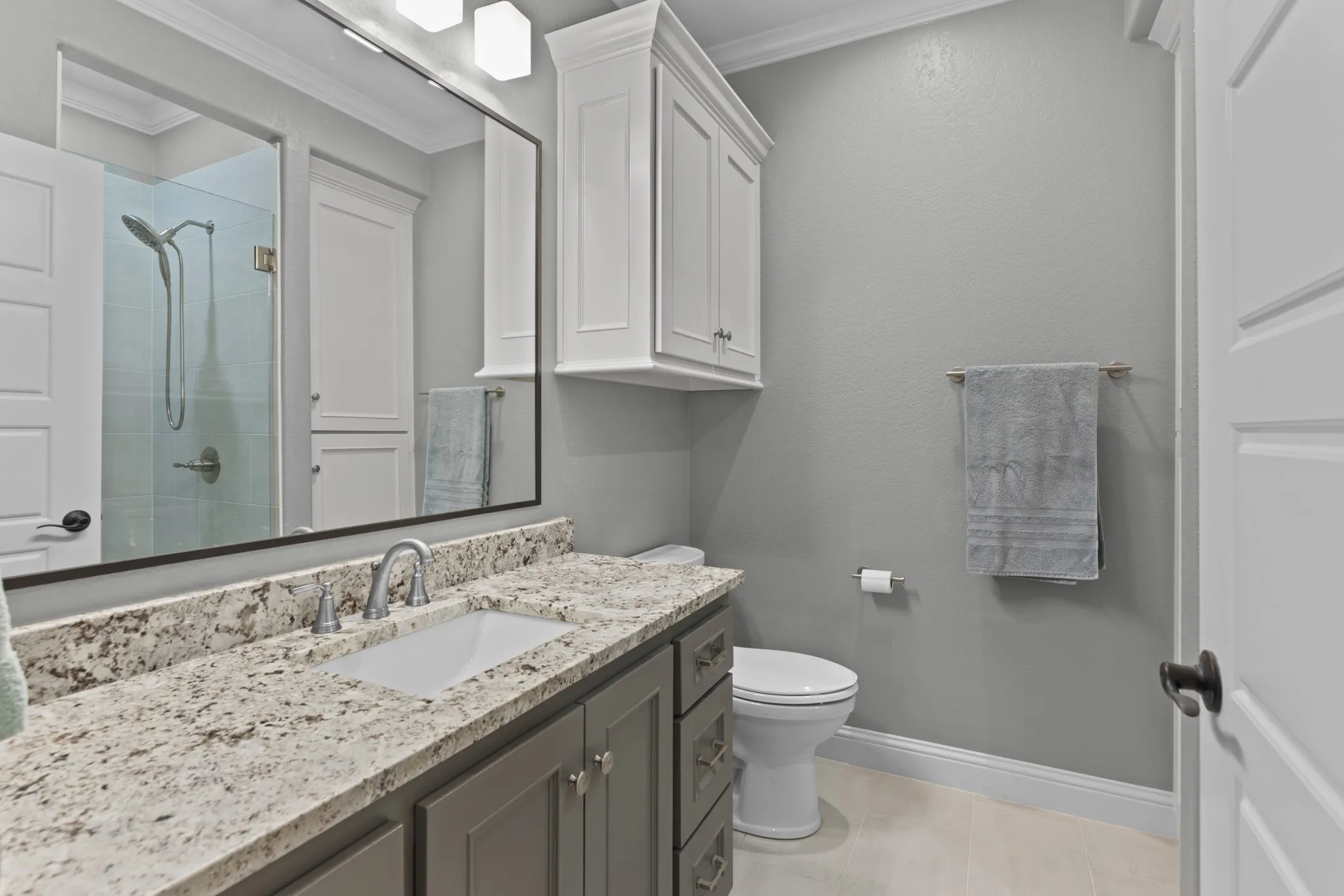 White shaker-style cabinetry with crown molding, a vanity with a white rectangular undermount sink, a stone-finish countertop, and a glass-enclosed shower with a rainfall showerhead
