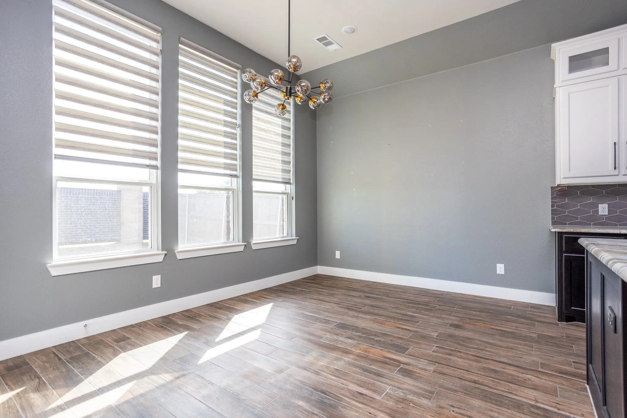 Triple windows with layered shades, wood-finish flooring, white cabinetry, dark gray backsplash, and a contemporary light fixture