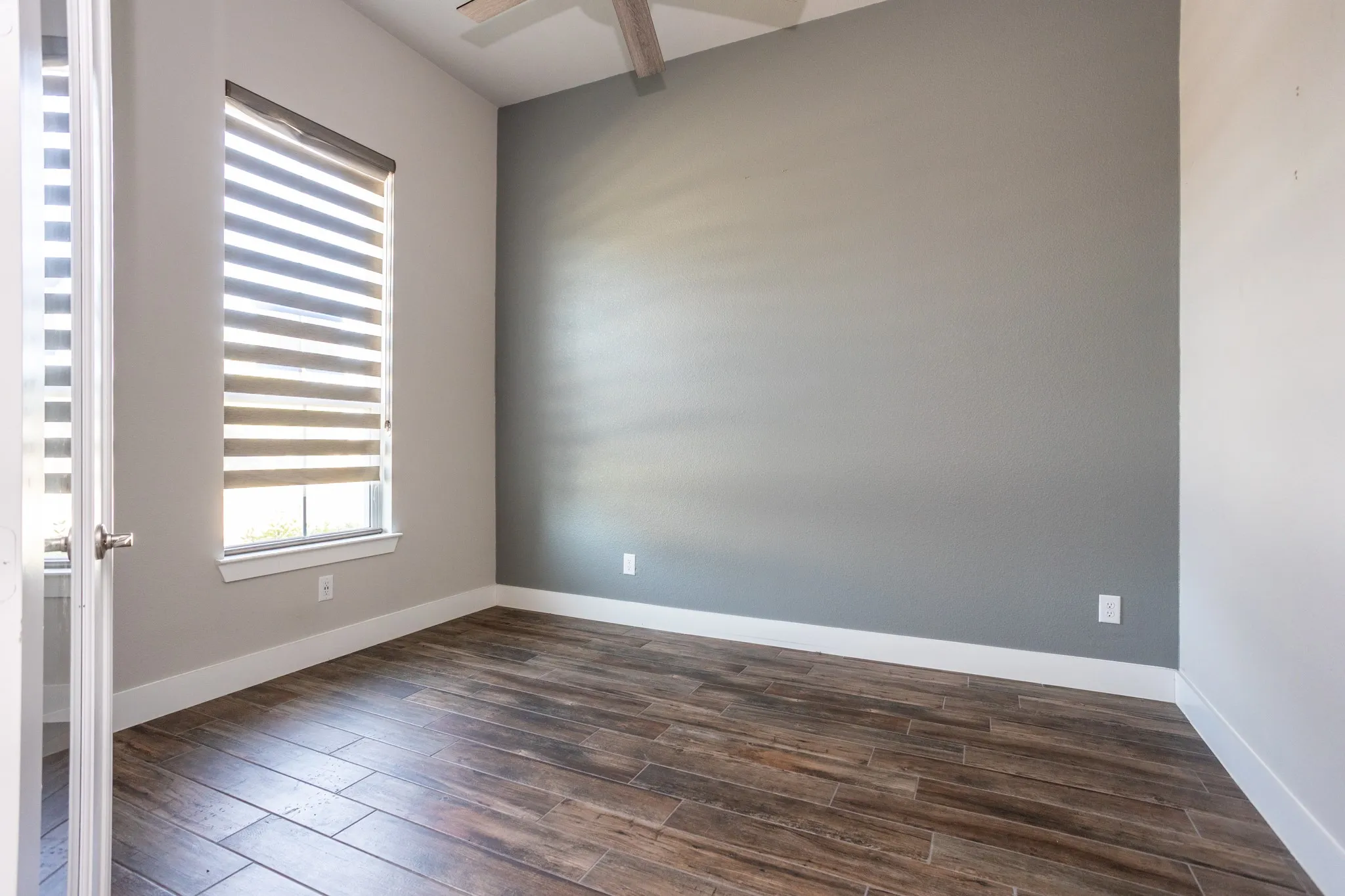 Wood-finish plank flooring, contemporary ceiling fan, accent wall, window with horizontal blinds, and crisp white trim