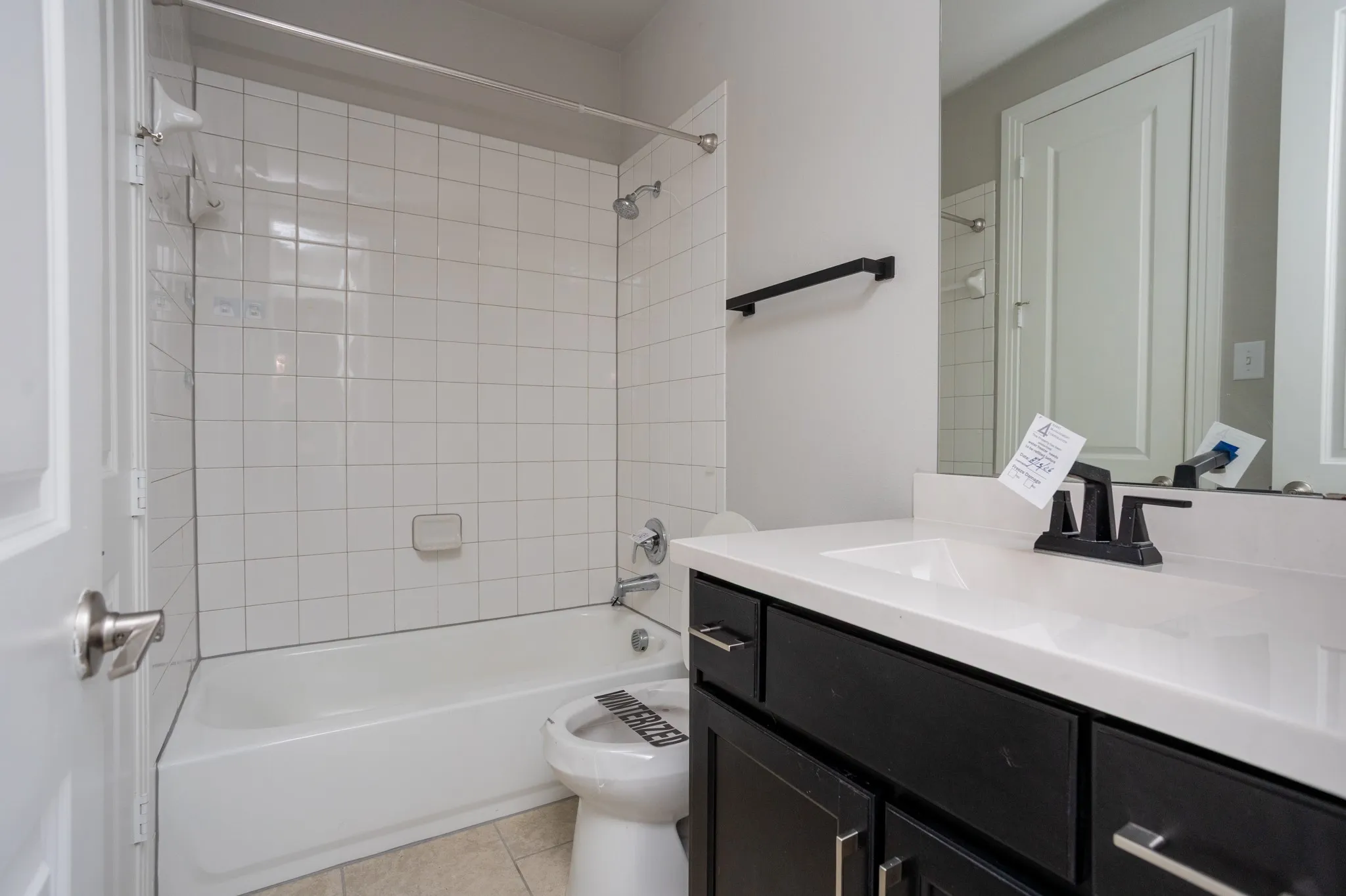 White tile bath-shower combination with chrome fixtures, dark wood-finish vanity with white countertop, and integrated rectangular sink