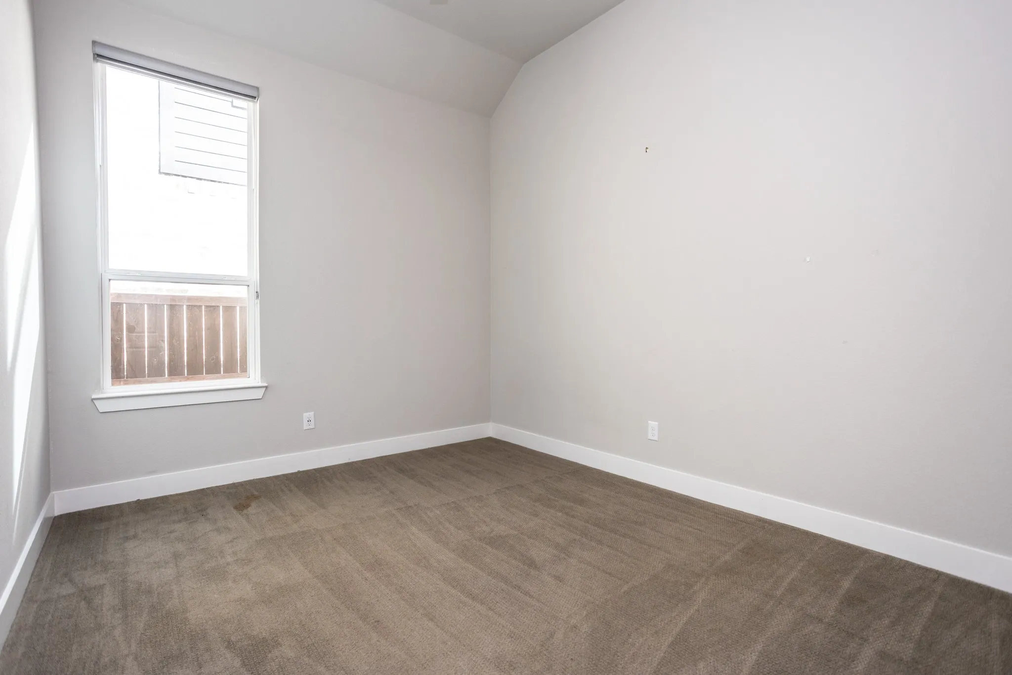 Neutral-toned walls with vaulted ceiling, plush carpet flooring, and a double-hung window with a window treatment