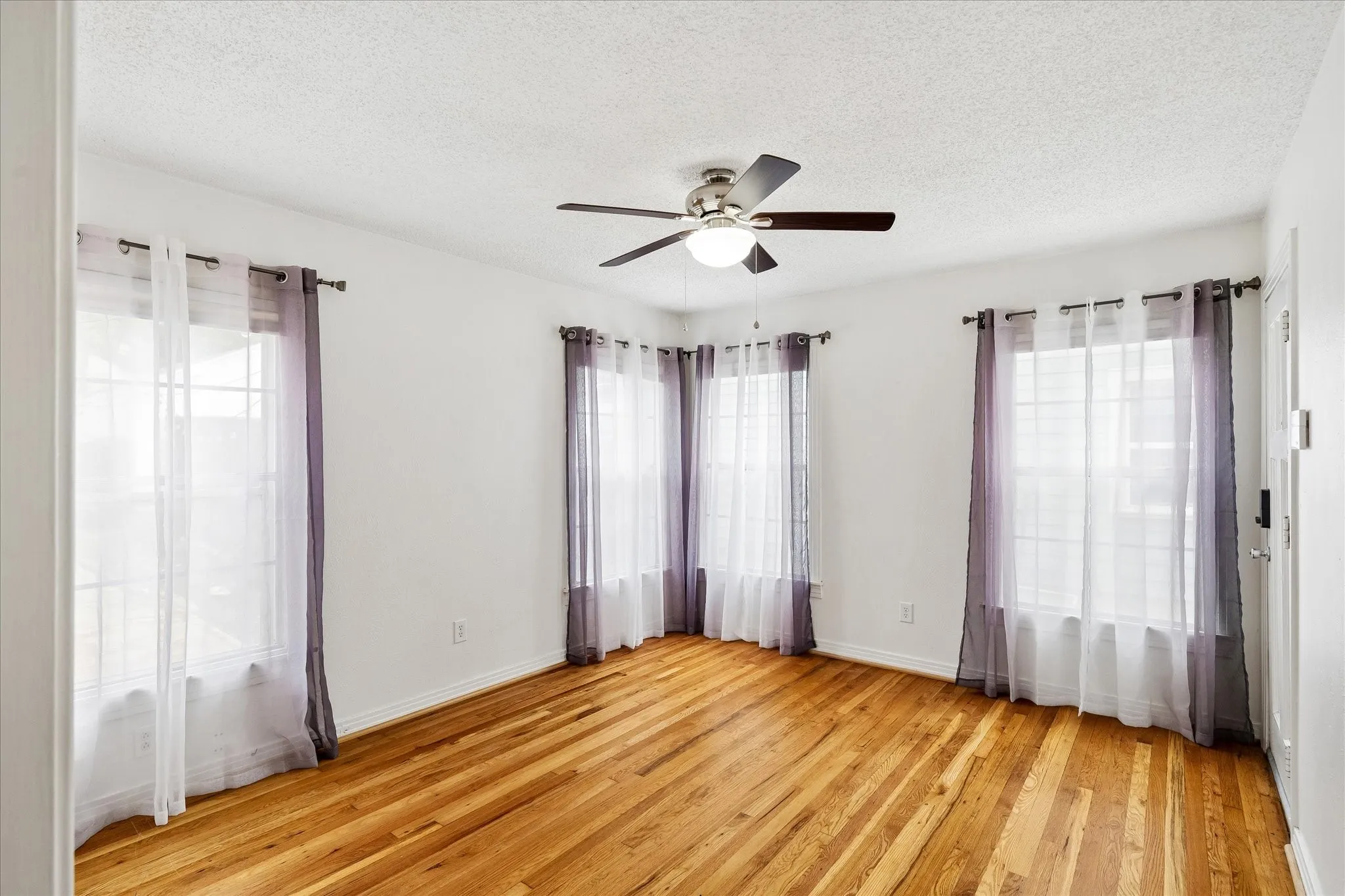 Hardwood flooring, white walls, multiple windows, ceiling fan with integrated lighting, and a paneled door