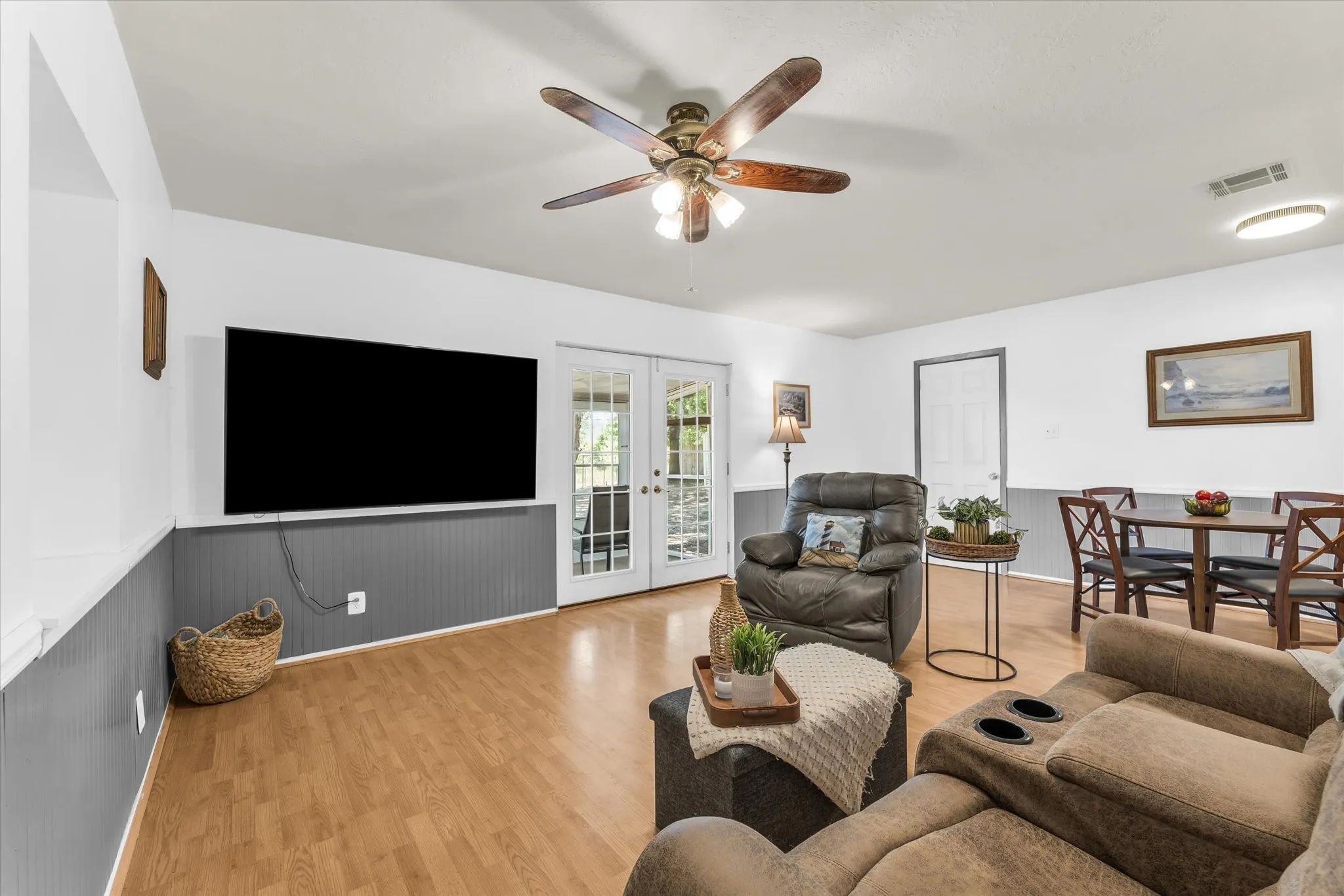 Spacious living area featuring wood-finish flooring, white walls with gray wainscoting, and a ceiling fan with integrated lighting