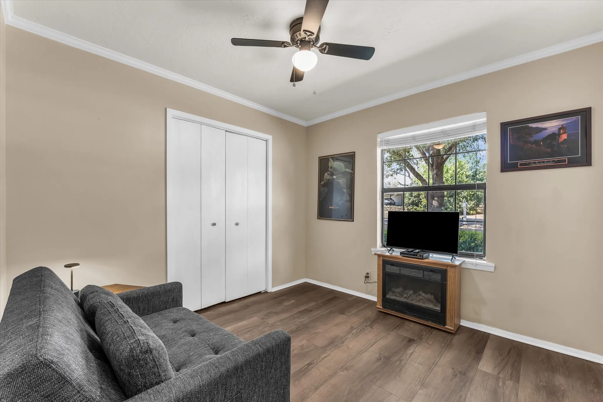 Wood-finish flooring, white bi-fold closet doors, ceiling fan with integrated lighting, white crown molding, and a window with exterior tree scenery