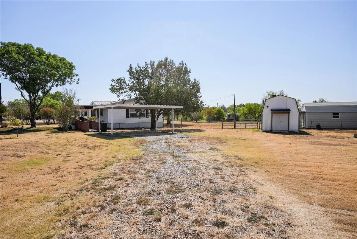 White single-story residence with a metal carport, mature trees, and a gravel driveway