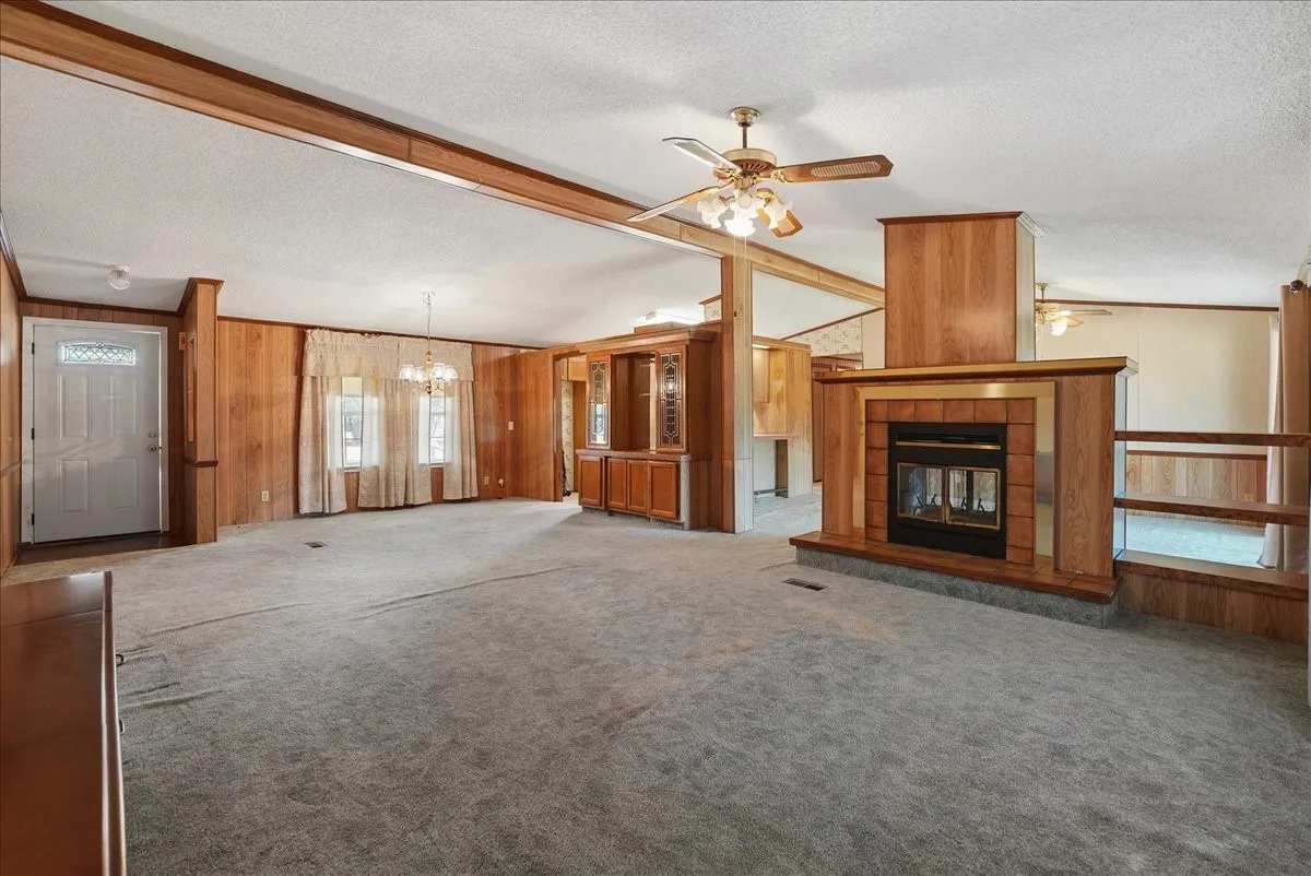 Spacious living area featuring a fireplace with brick and wood surround, wood-paneled walls, built-in cabinetry, and a ceiling fan