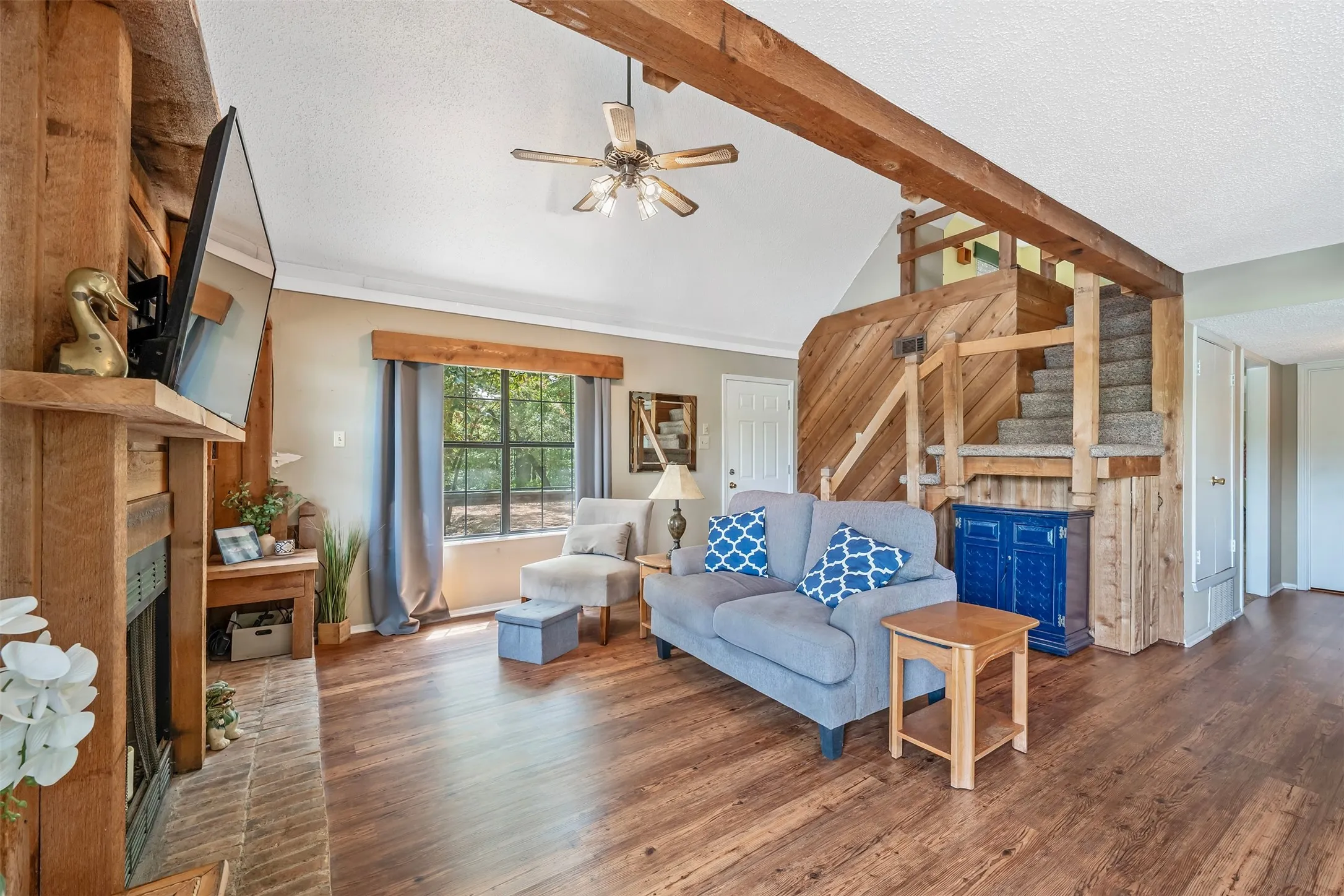 Vaulted ceiling with exposed wood beams, wood-finish flooring, built-in fireplace with brick surround, built-in wooden staircase, and ceiling fan with integrated lighting