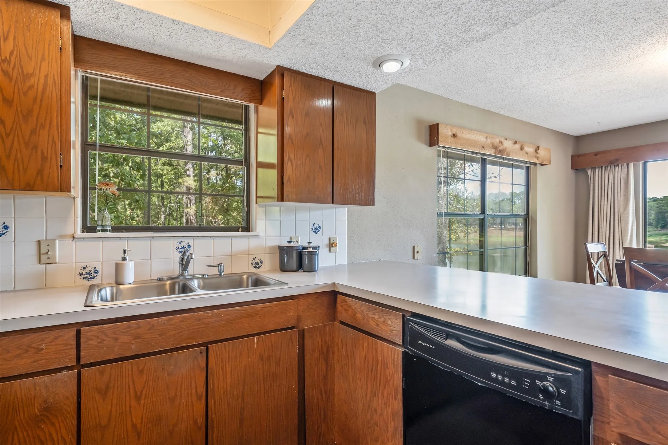 Kitchen featuring wood cabinetry, a double basin stainless steel sink, a dishwasher, a white tile backsplash, and light-toned countertops