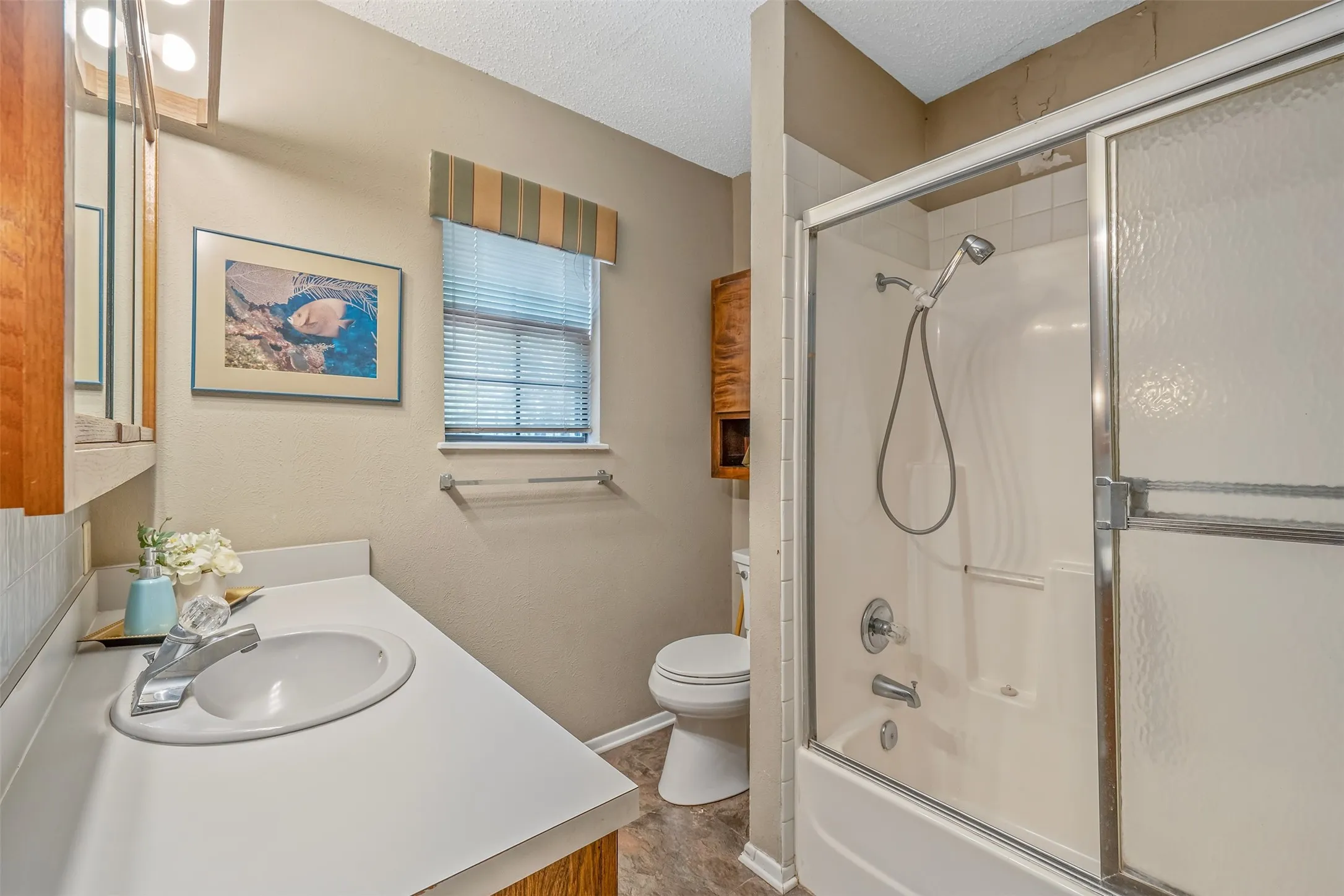 White vanity with integrated sink and chrome faucet, wood-finish cabinetry, shower/tub combination with sliding glass doors, white toilet, and window with blinds