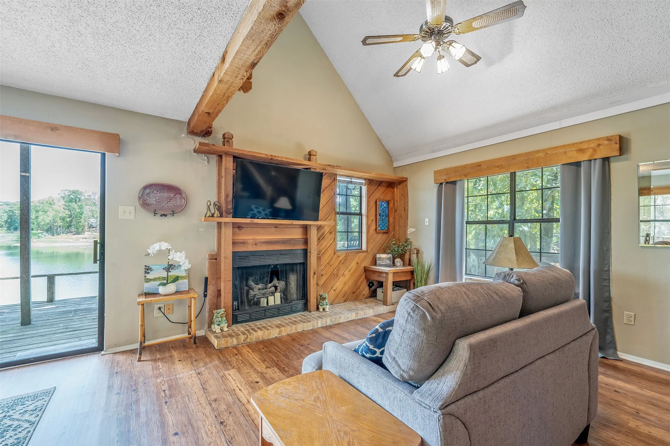 Vaulted ceiling with exposed wood beam, wood-finish flooring, brick fireplace with wood surround, sliding glass door to exterior decking, and a ceiling fan with integrated lighting