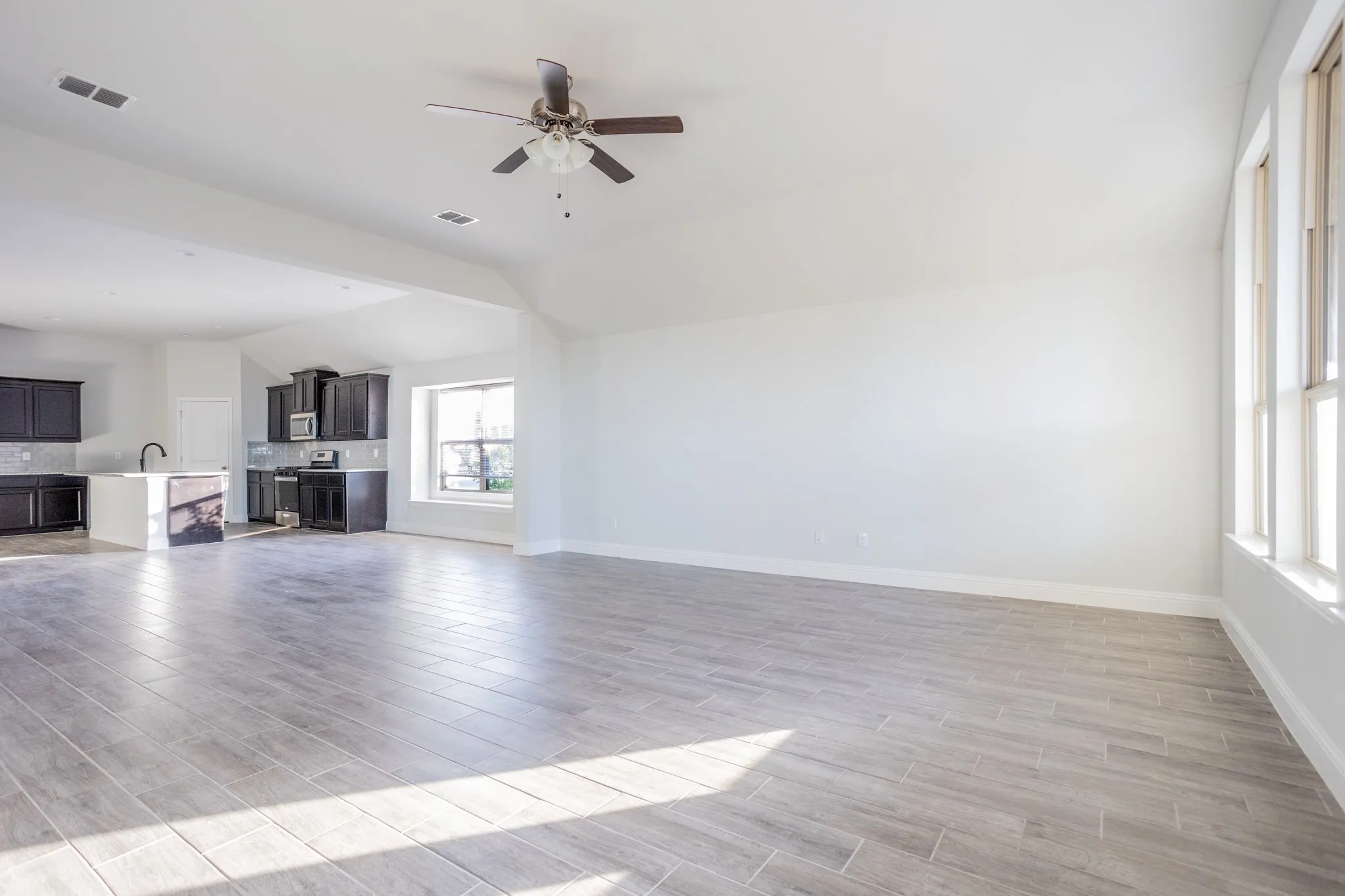 Spacious open-concept living area with wood-finish flooring throughout, abundant natural light from large windows, ceiling fan with integrated lighting, and adjacent kitchen with cabinetry and appliances visible in the background