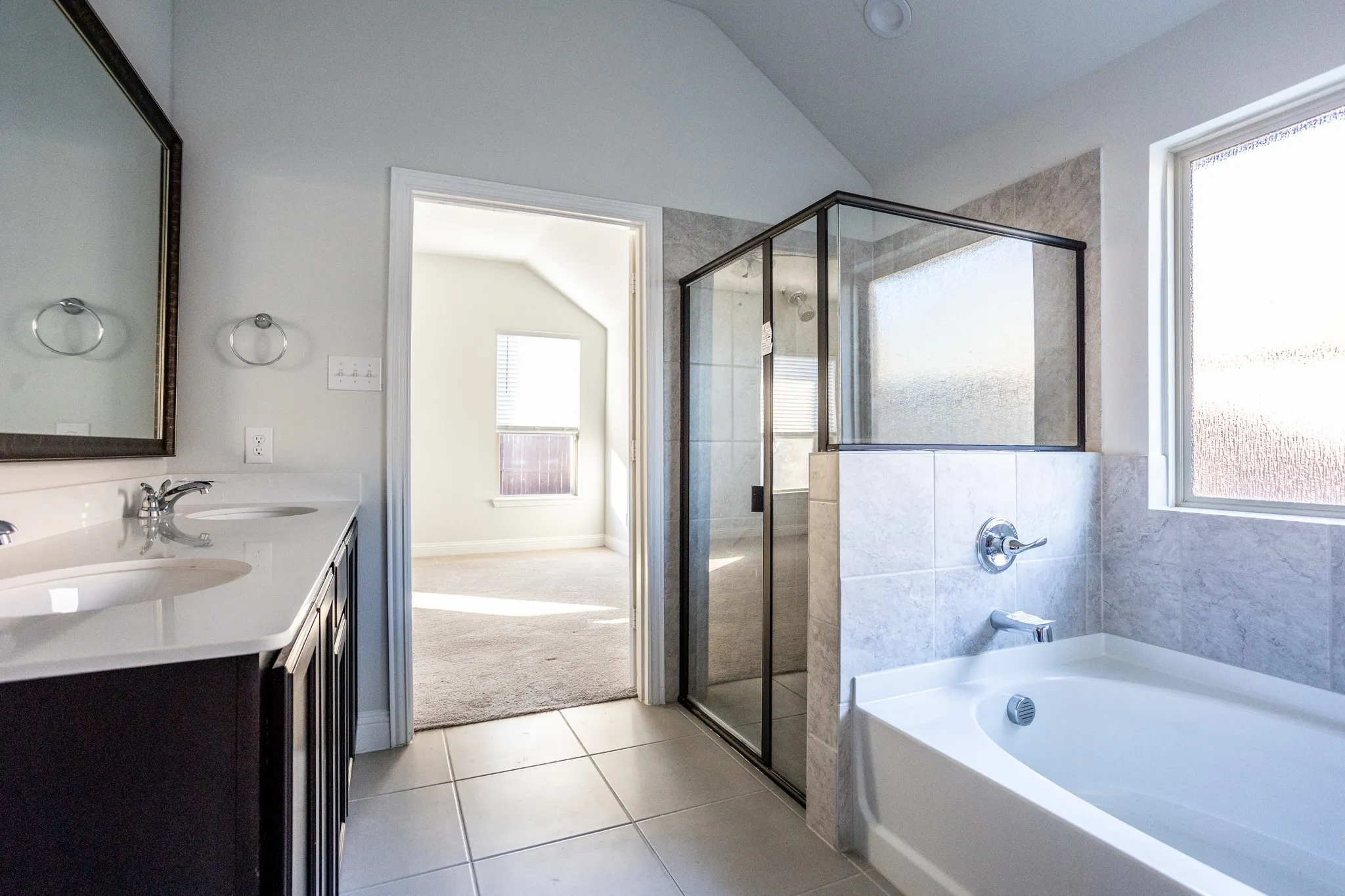 White tile flooring, dual sink vanity with white countertop, chrome fixtures, glass-enclosed shower, and built-in soaking tub