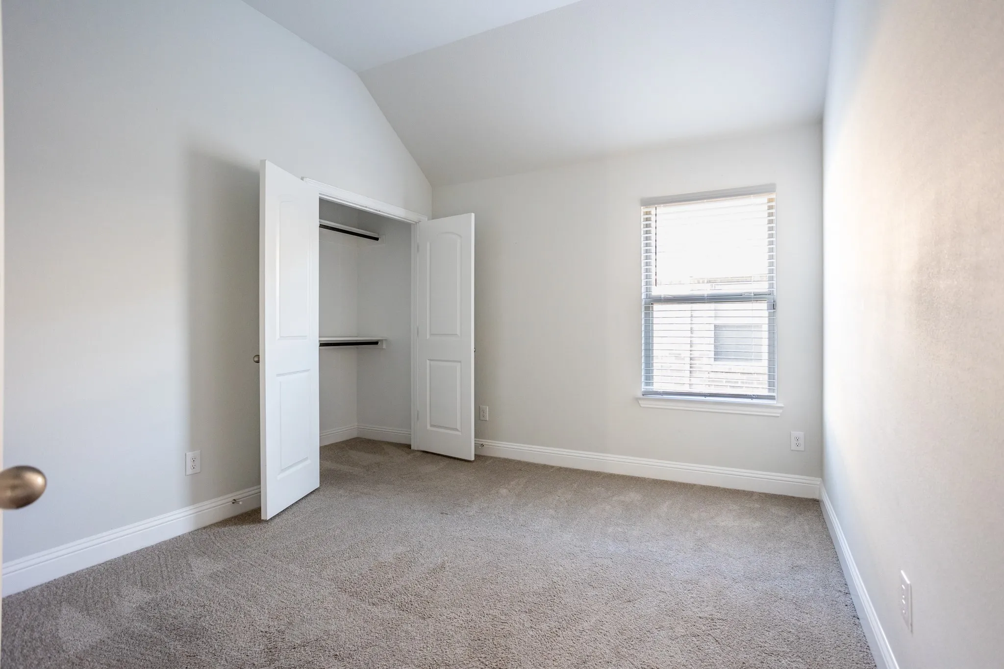 Vaulted ceiling with neutral paint, carpeted flooring, window with blinds, and bi-fold closet doors with built-in shelving