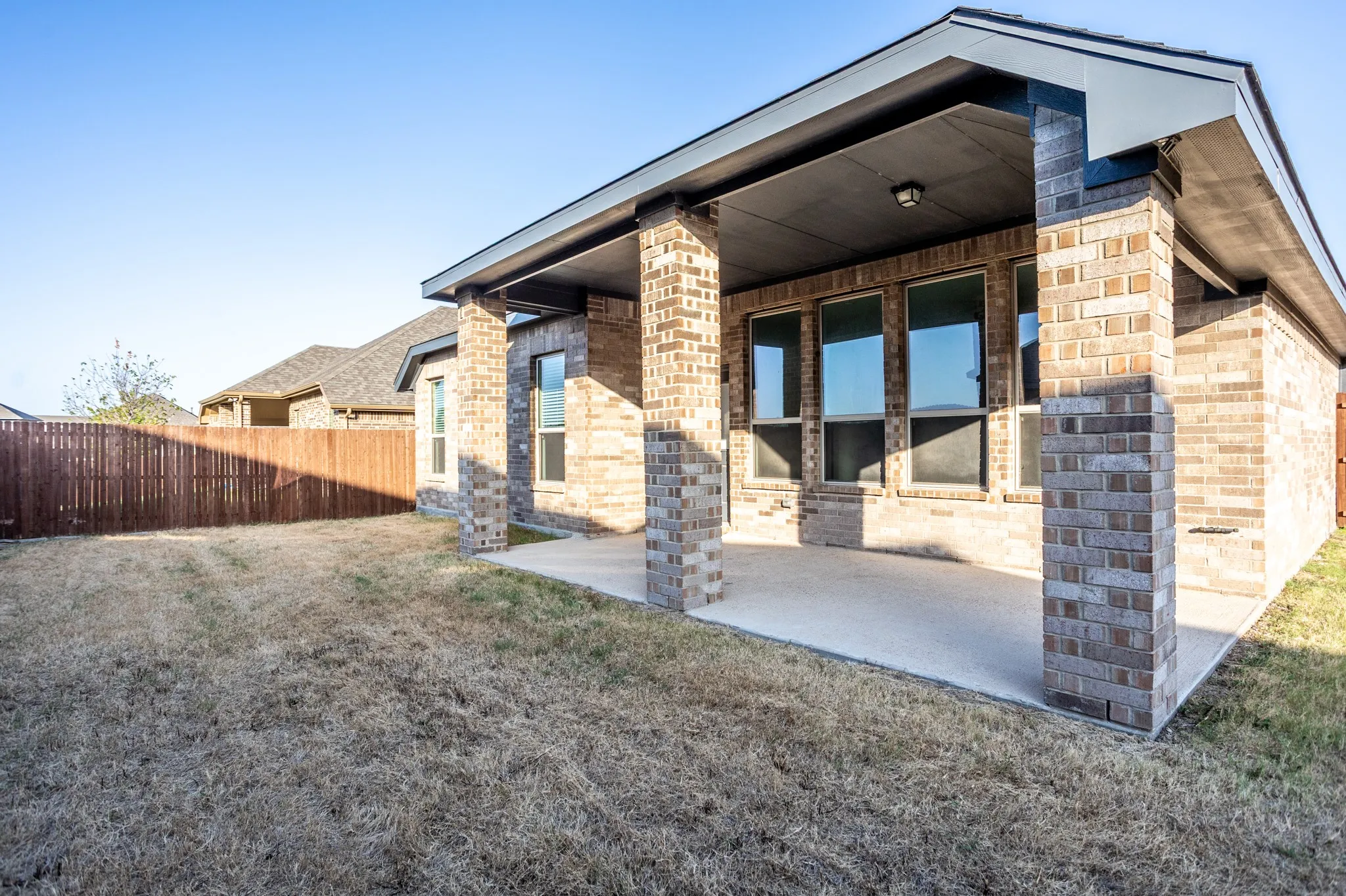 Modern brick and stone exterior with covered patio featuring multiple large windows, metal support columns, and expansive concrete entryway