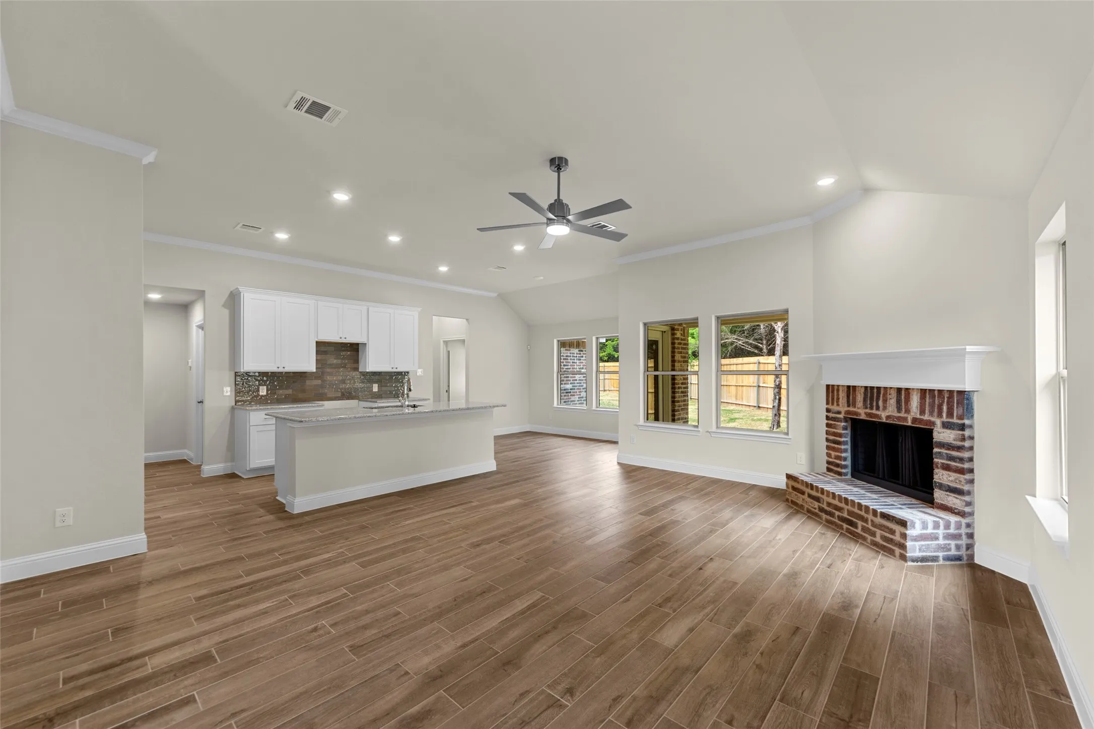 Expansive living area featuring wood-finish flooring, a brick fireplace with a white mantel, and vaulted ceilings with a ceiling fan