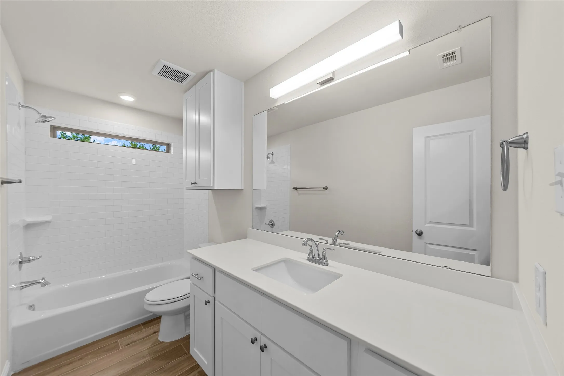 White subway tile tub surround, elongated vanity mirror, white cabinetry, wood-finish flooring, and white solid surface countertop