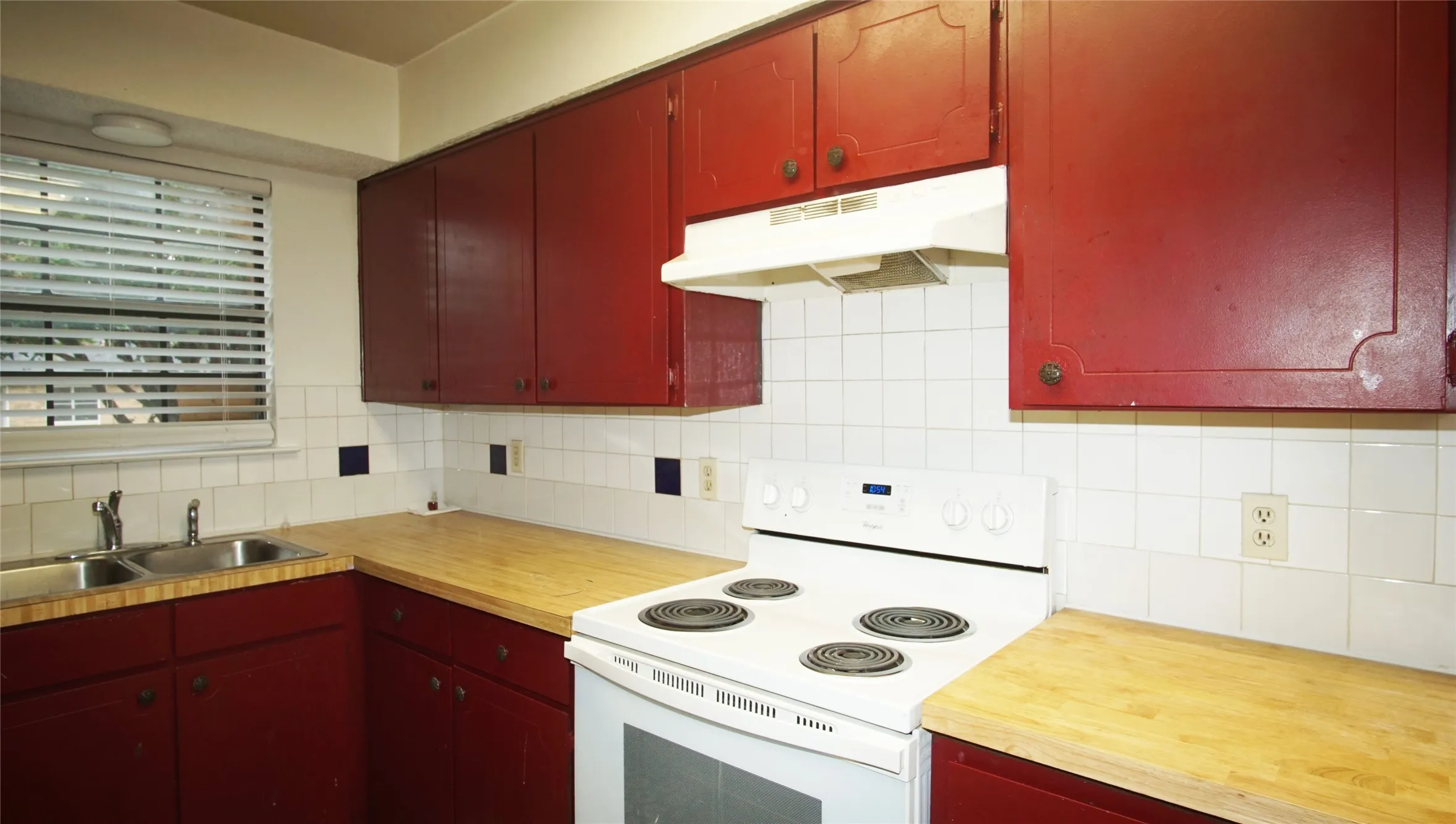 Kitchen with white electric stove, red cabinets, tasteful backsplash, under cabinet range hood, and wooden counters