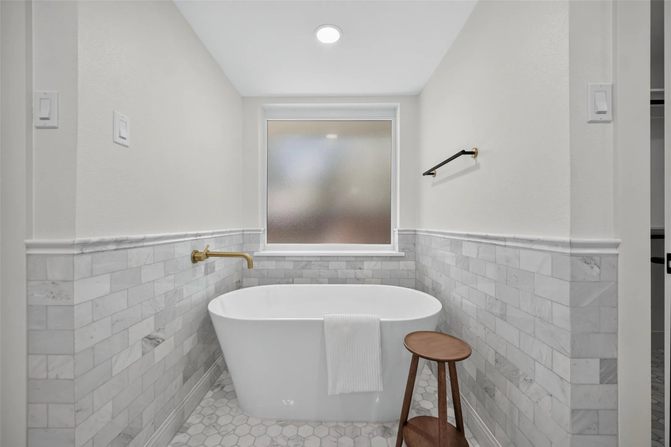 Freestanding soaking tub with a wall-mounted brass faucet, subway tile wainscoting, hexagonal floor tiles, and a frosted window