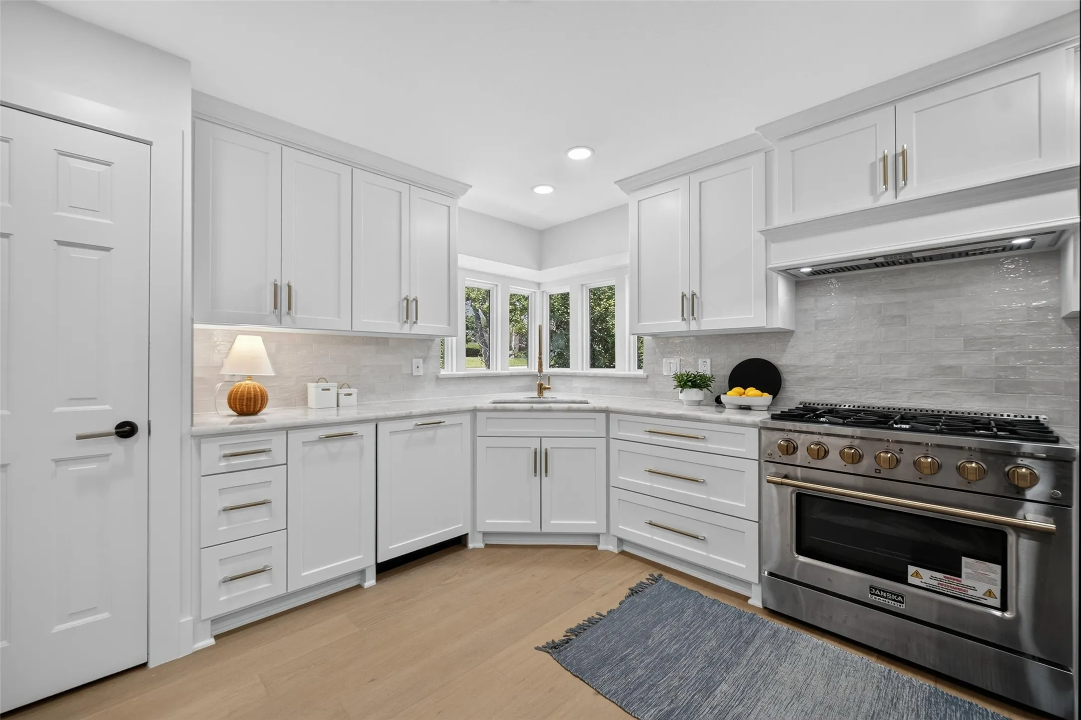 White shaker cabinetry with crown molding, stainless steel range with matching hood, light wood-finish flooring, undermount sink, and tiled backsplash