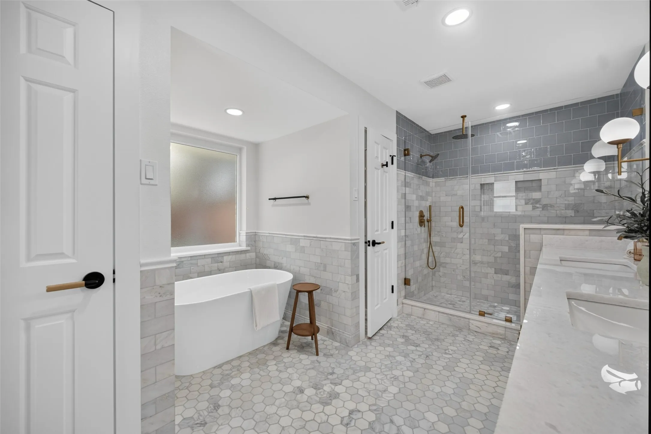 Spa-inspired primary bathroom featuring a freestanding soaking tub with wooden accent stool, walk-in shower with frameless glass enclosure and subway tile surround, hexagonal tile flooring, floating vanity with integrated sink, recessed and pendant lighting, and dual-sink configuration with chrome fixtures