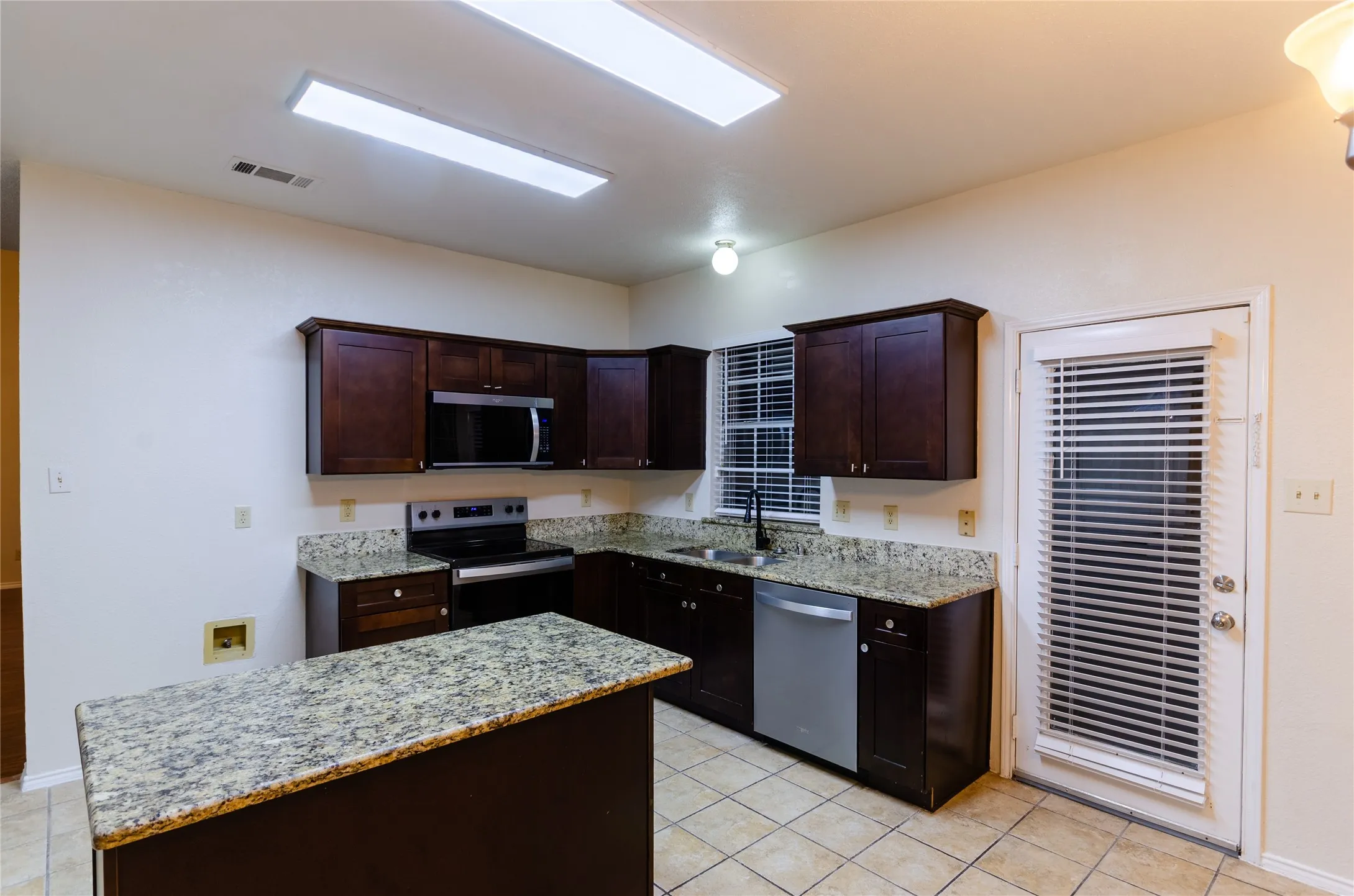 Kitchen featuring dark wood-finish cabinetry, light speckled countertops, a center island with a matching countertop, a stainless steel dishwasher, and tile flooring