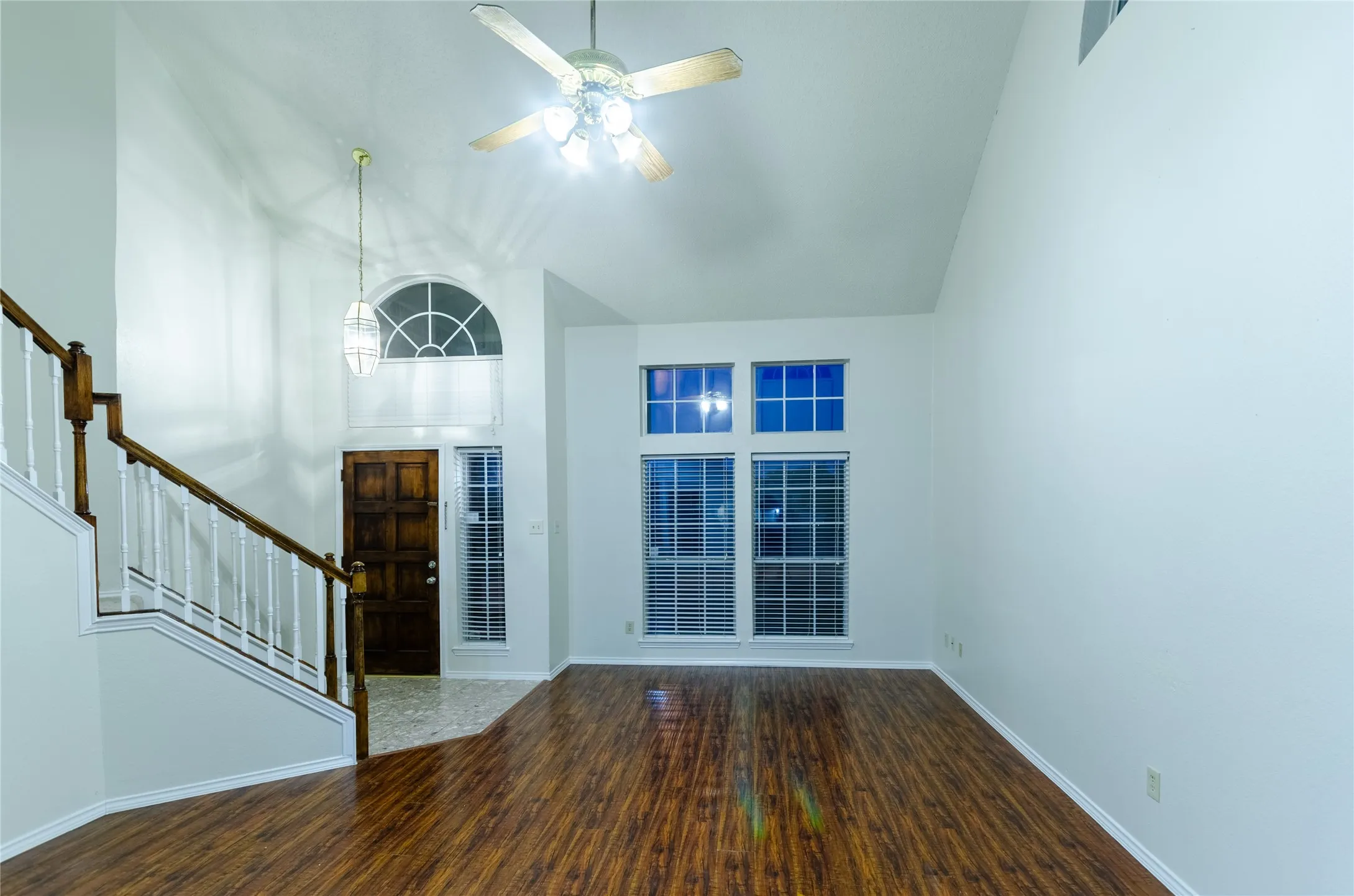 Wood-finish flooring throughout, prominent staircase with wood handrail, high ceilings, ceiling fan with integrated lighting, and multiple windows with blinds