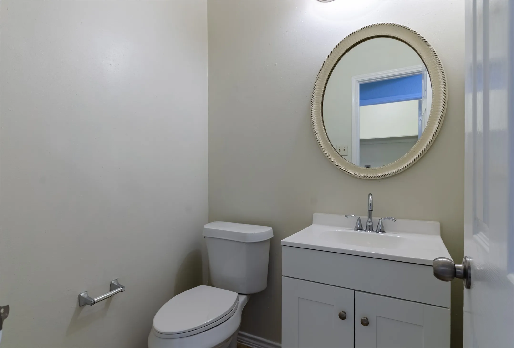 Half bath featuring a white ceramic toilet, vanity sink with brushed nickel faucet and hardware, soft-close cabinetry with ample storage, round beaded mirror with polished frame, and neutral wall finishes