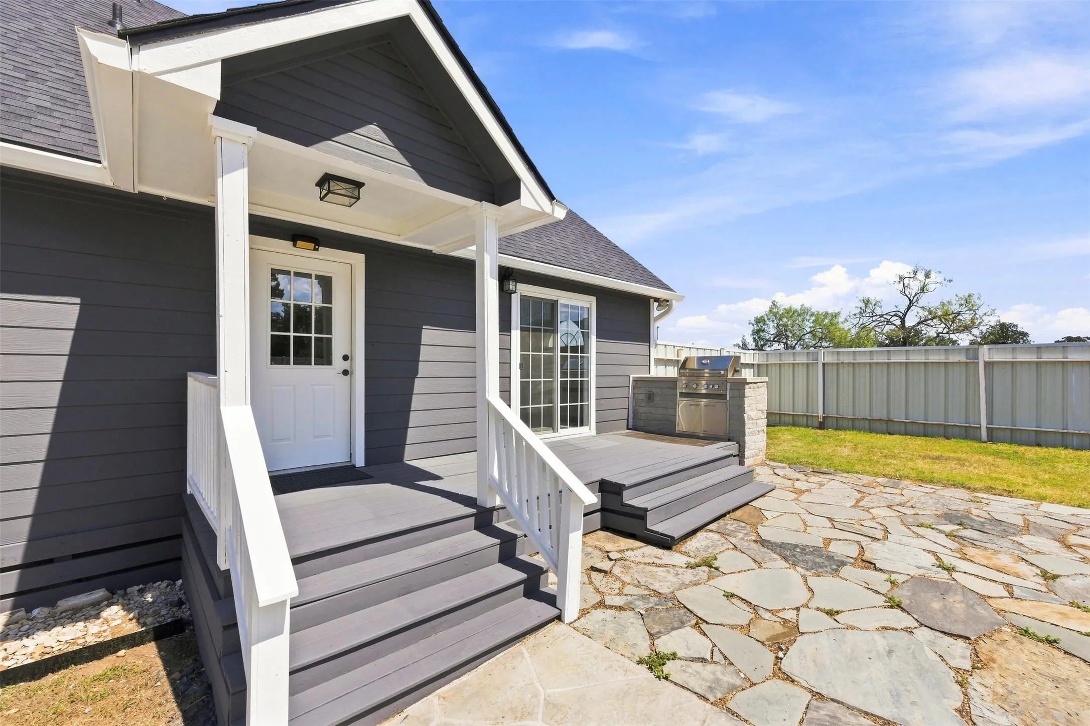 Exterior featuring horizontal siding, covered porch with white railings, multi-pane entry door, and a paver patio with built-in grill station