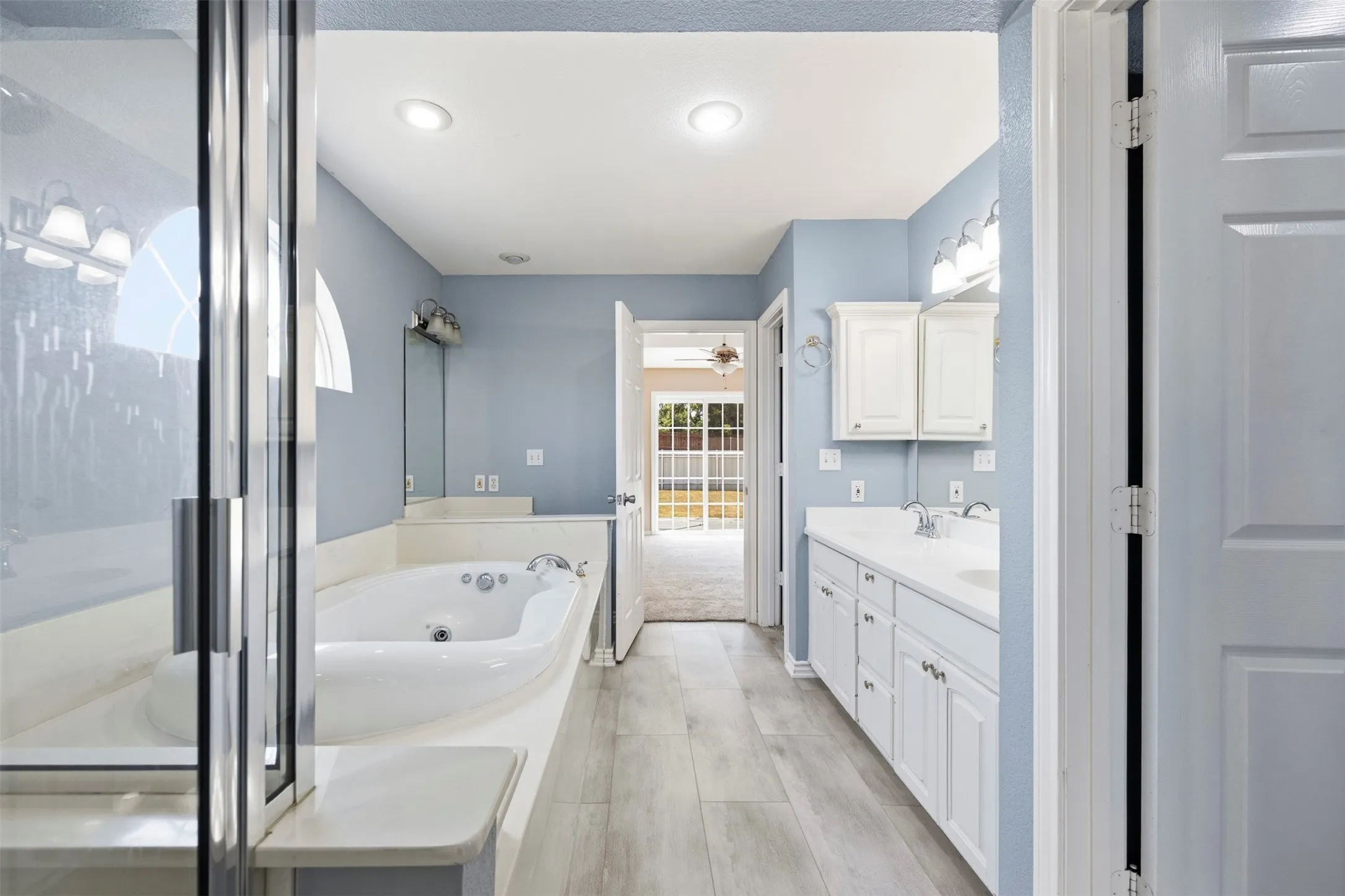 Elongated soaking tub, separate glass-enclosed shower, dual vanity with white cabinetry, and wood-finish flooring