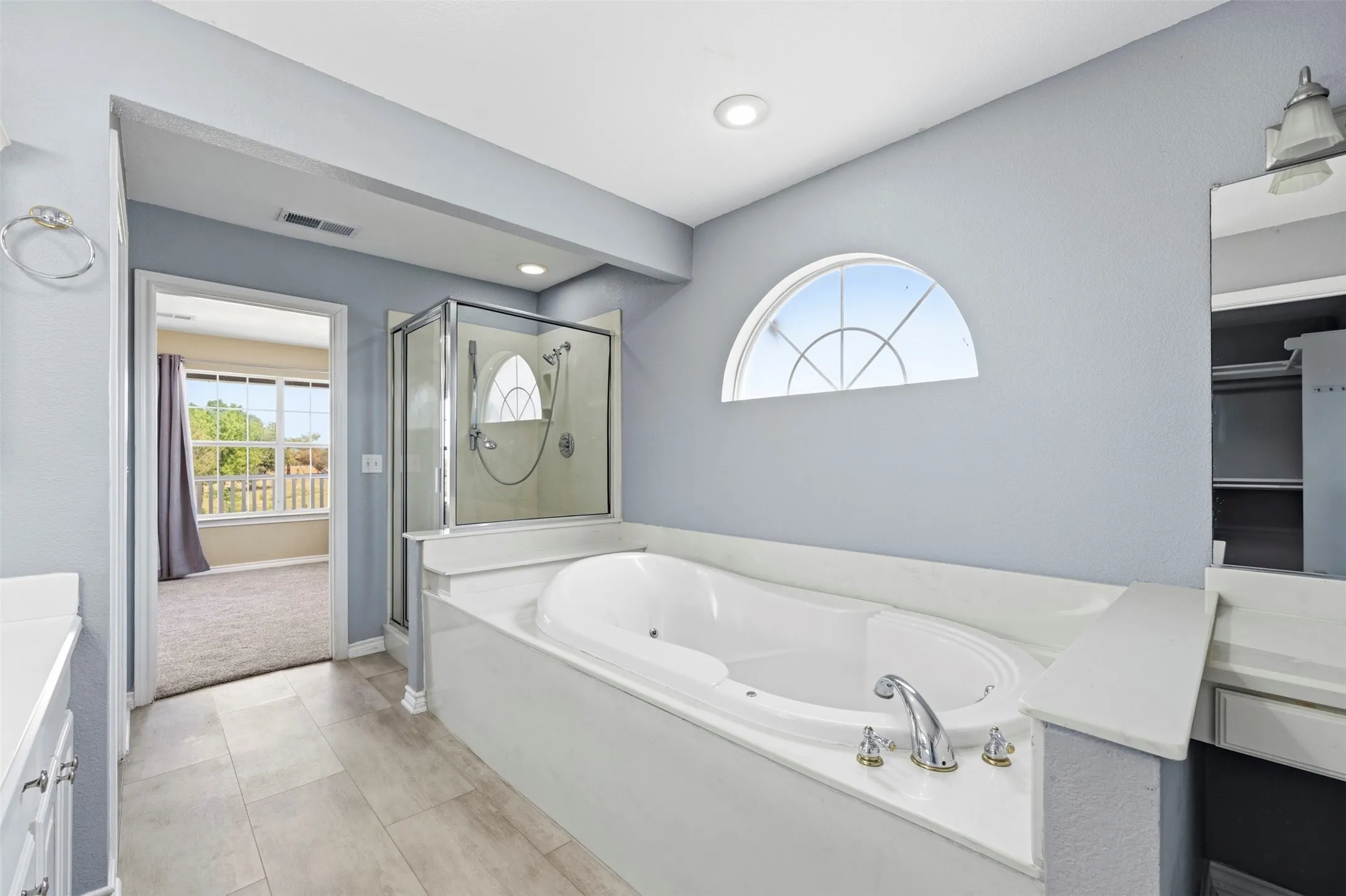 Soaking tub with deck-mounted fixtures, glass-enclosed shower, arched window, tile flooring, and integrated vanity cabinetry