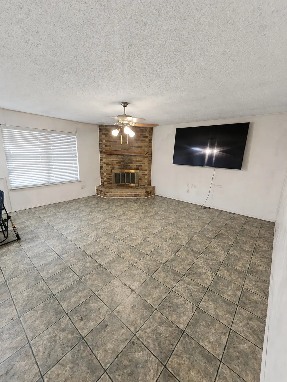 Spacious room featuring a brick fireplace, ceiling fan with integrated lighting, window with horizontal blinds, and tile flooring