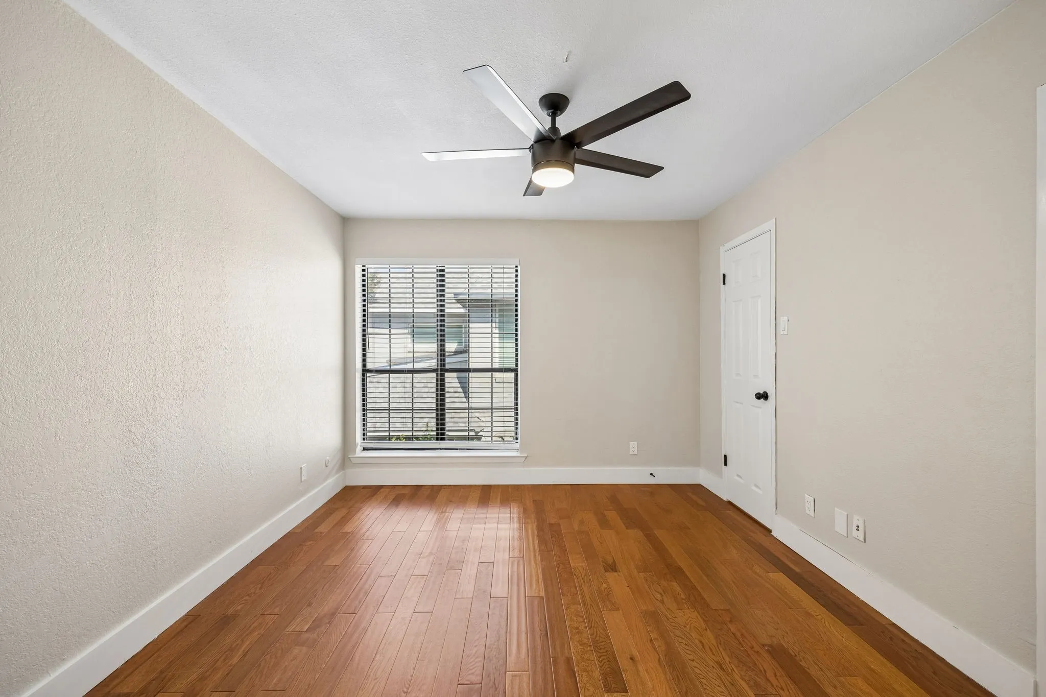 Spacious room featuring wood-finish flooring, a contemporary ceiling fan with integrated lighting, a window with horizontal blinds, and a white paneled door with black hardware