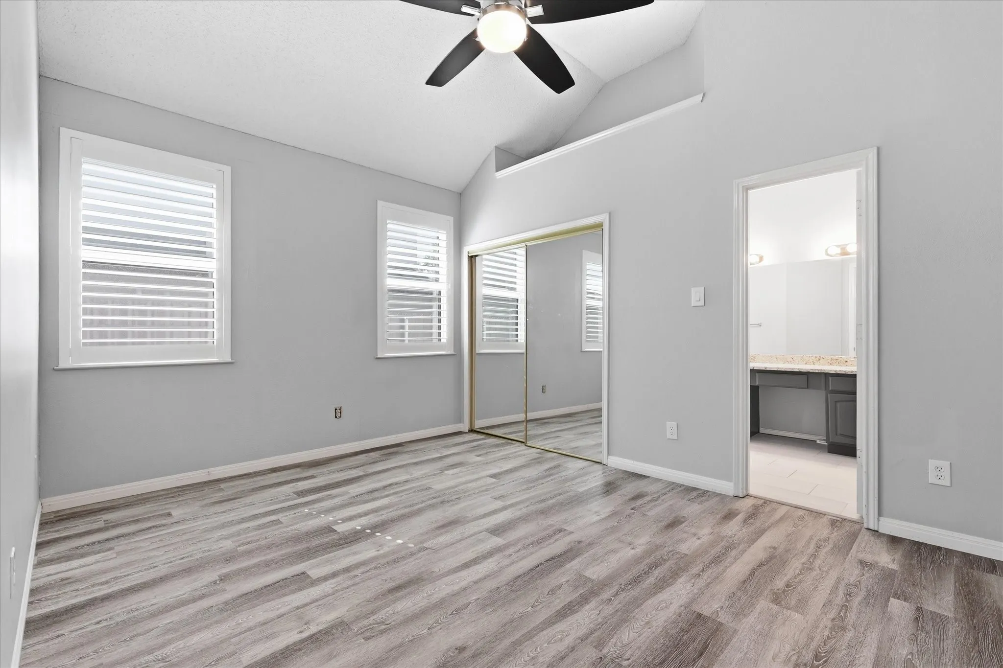Vaulted ceiling, wood-finish flooring, two windows with plantation shutters, mirrored closet doors, and an attached bathroom with a double vanity