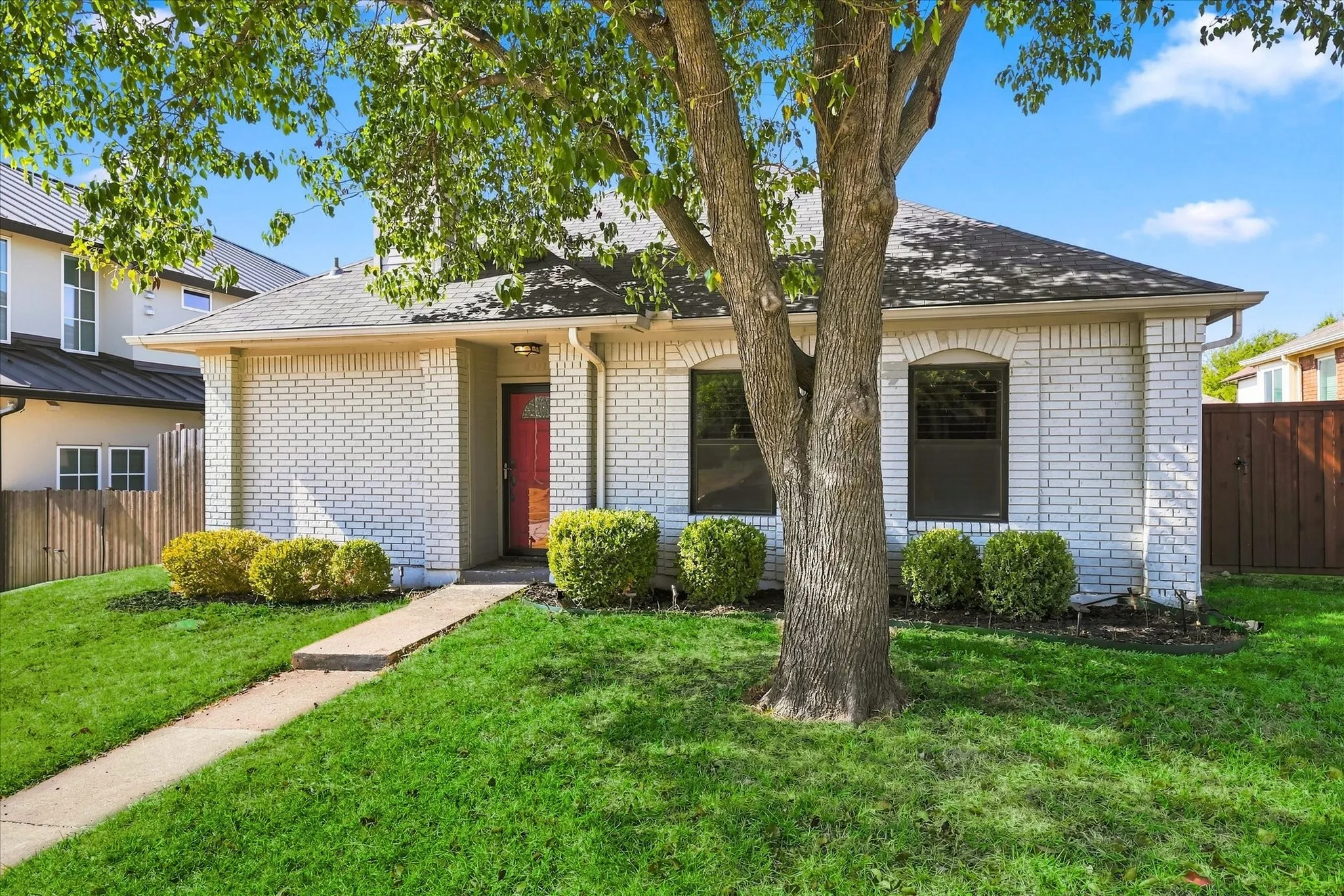 White brick exterior with a red entry door, framed by established shrubbery and a mature tree