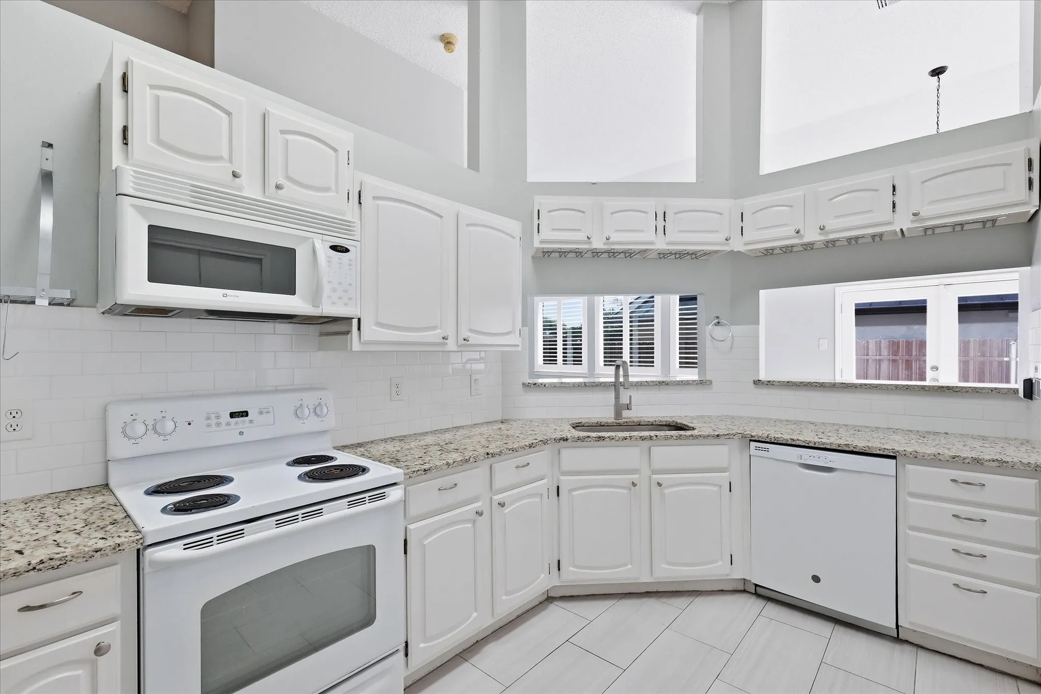 White cabinetry with curved panel doors, granite countertops, white subway tile backsplash, white built-in appliances, and light-toned tile flooring