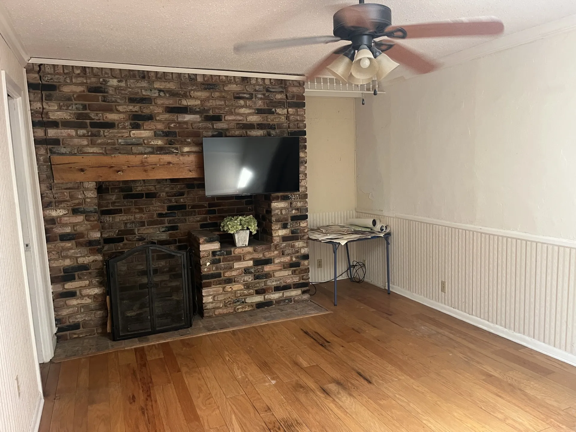 Brick fireplace with wood mantel, built-in shelving, wood-finish flooring, ceiling fan, and wainscoting
