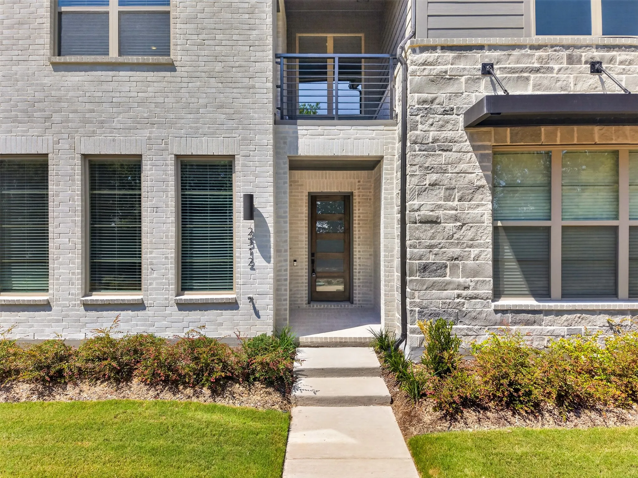Exterior featuring light brick, natural stone accents, a modern entry door with glass panels, a second-story balcony with metal railings, and a streamlined awning