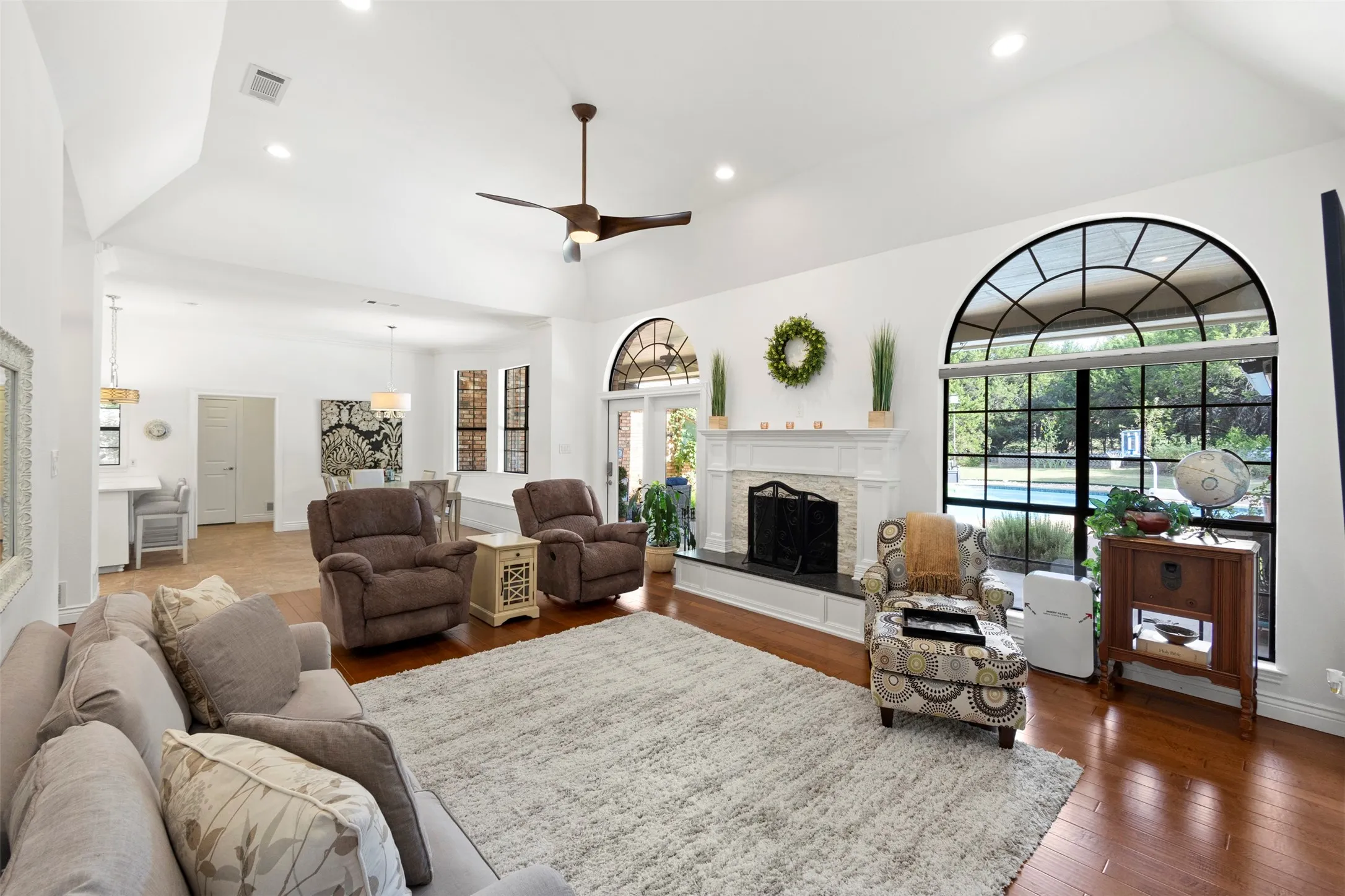 Vaulted ceiling with recessed lighting, wood-finish flooring, white fireplace with dark hearth, and a large arched window with grid details