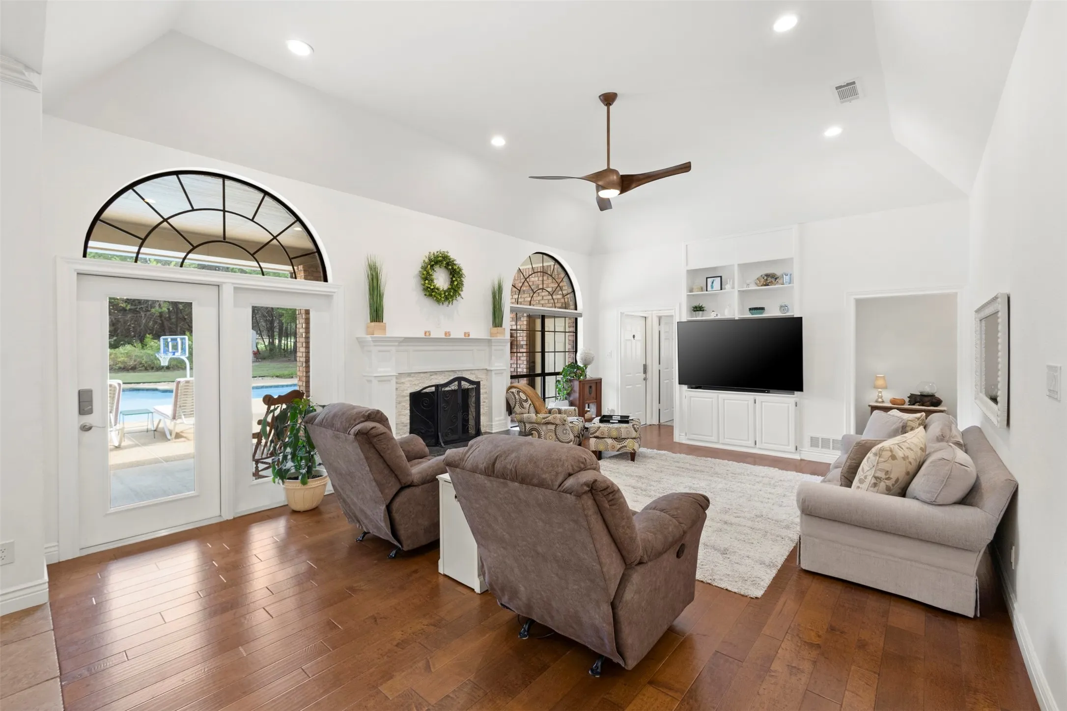 Vaulted ceiling with recessed lighting, wood-finish flooring, white fireplace with a stone surround, built-in cabinetry, and transom windows
