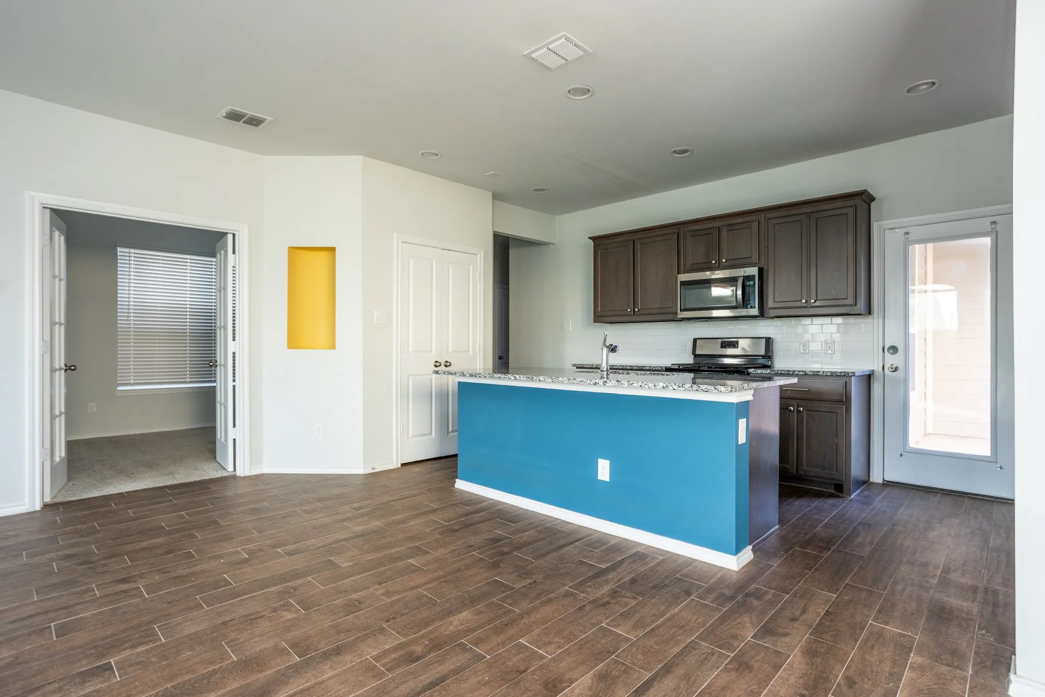 Open-concept living space with wood-finish flooring, a kitchen island, dark wood cabinetry, stainless steel appliances, and subway tile backsplash