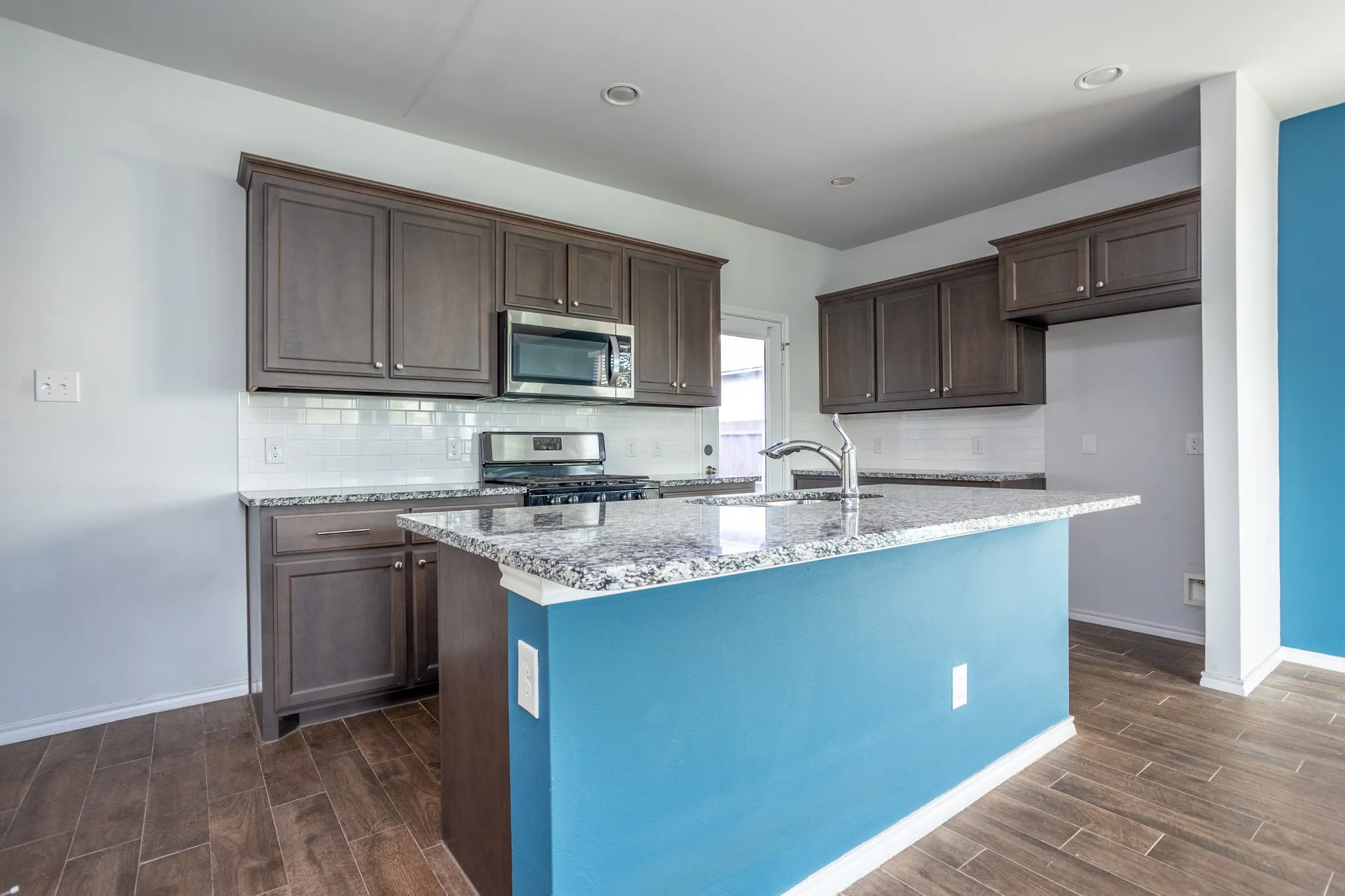 Kitchen featuring wood-finish flooring, dark wood cabinetry, white subway tile backsplash, and a central island with a granite countertop and sink