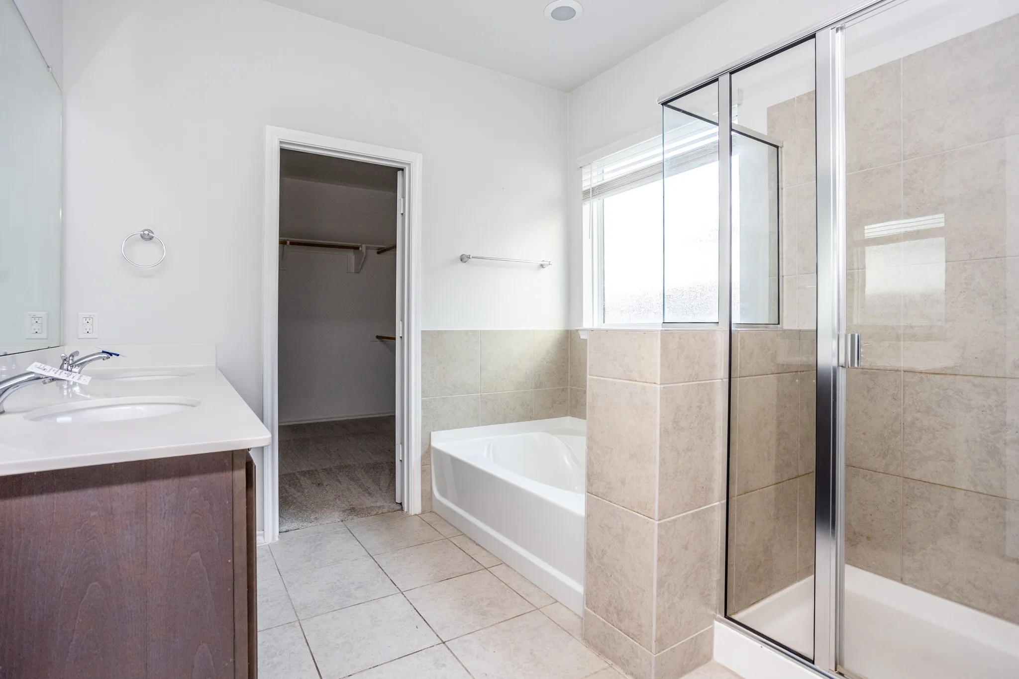 Dual-sink vanity with a light-toned countertop, tiled flooring, built-in bathtub, and a separate glass-enclosed shower