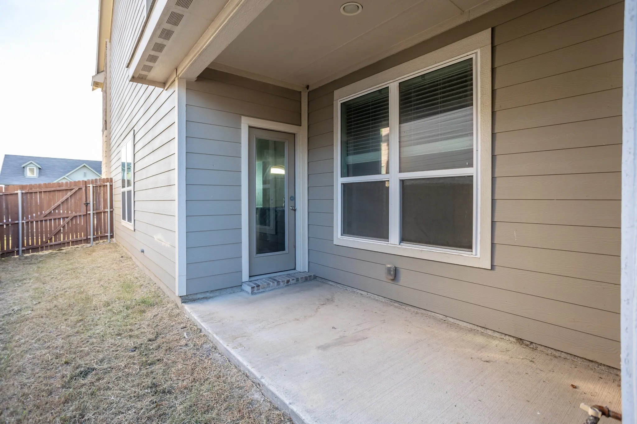Covered patio with concrete flooring and glass sliding door entry, framed by fiber cement siding in a neutral gray tone
