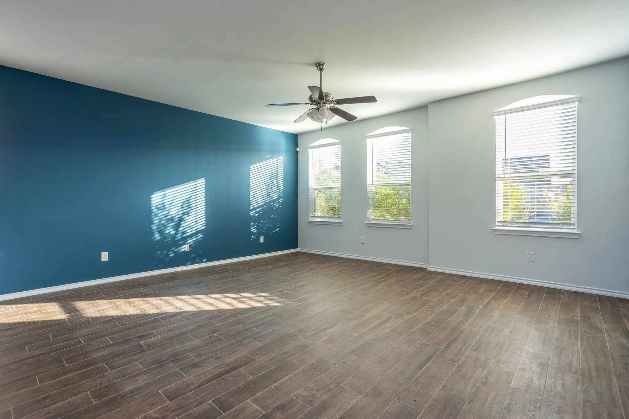 Spacious room featuring wood-finish flooring, a ceiling fan, and multiple arched windows with blinds