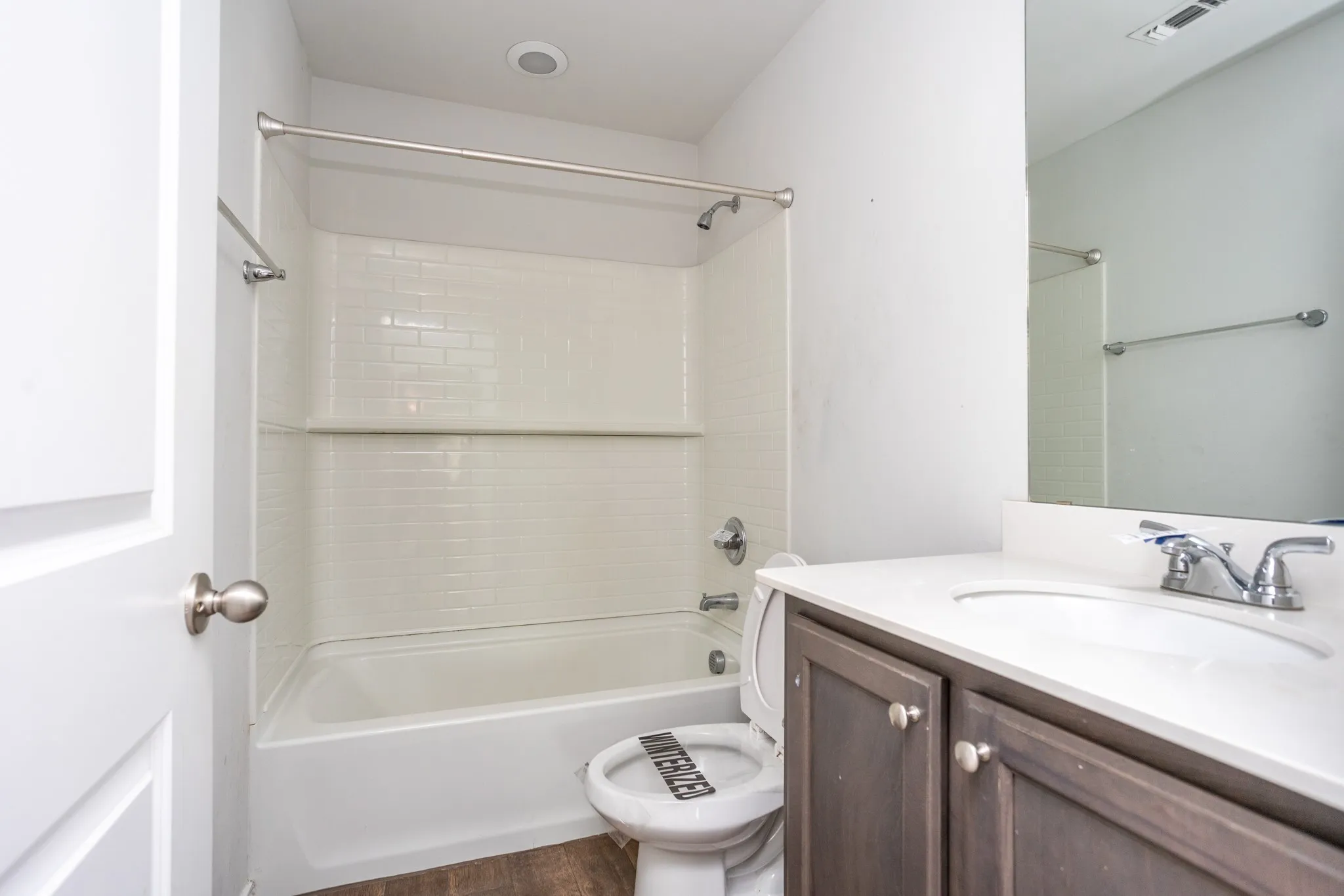 Bathroom featuring a vanity with a white countertop and wood-finish cabinetry, a full-size mirror, a shower-tub combination with subway tile surround, and wood-finish flooring