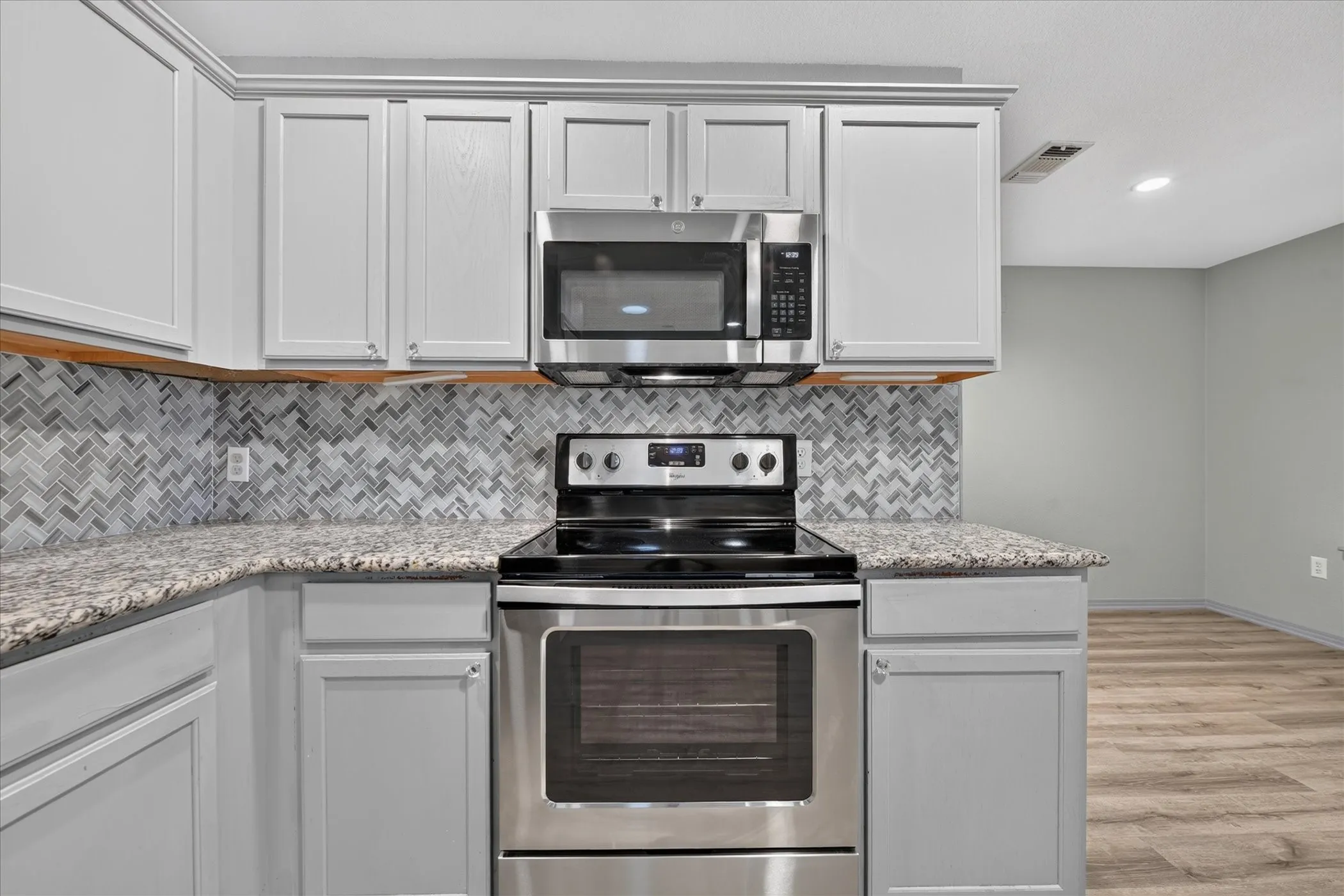 Stainless steel range and microwave, light-toned cabinetry, herringbone-patterned tile backsplash, speckled countertops, and wood-finish flooring