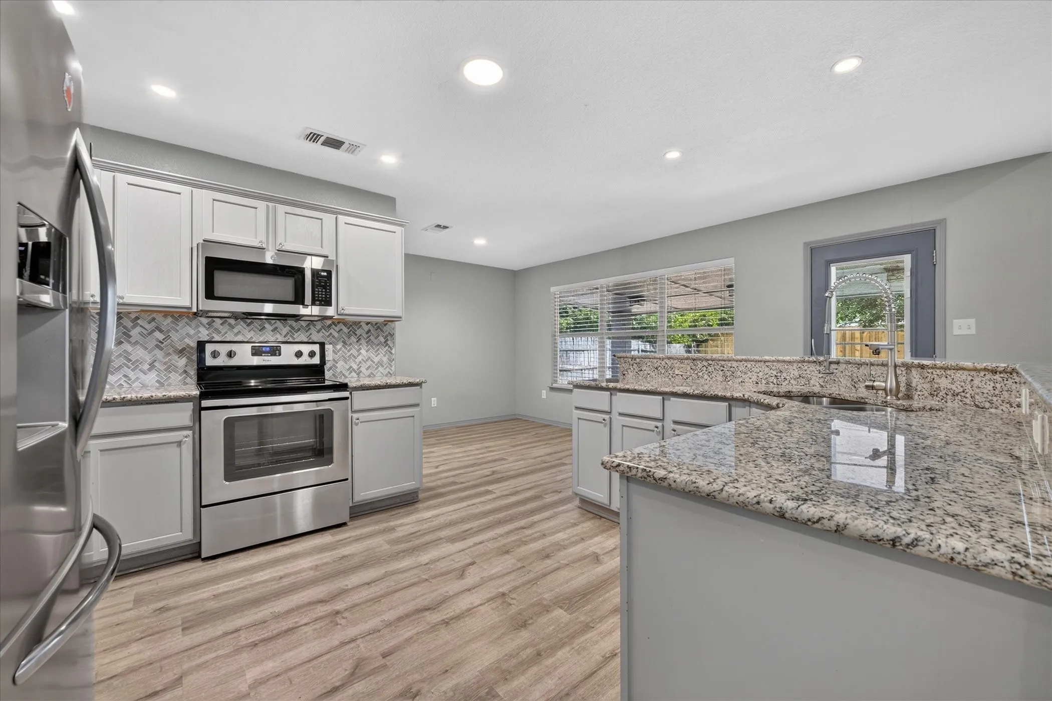 Modern kitchen featuring stainless steel appliances, white cabinetry with ample storage, granite countertops, a center island with breakfast bar seating, recessed ceiling lighting, wood-finish flooring, and abundant natural light from multiple windows and glass doors overlooking the outdoor space